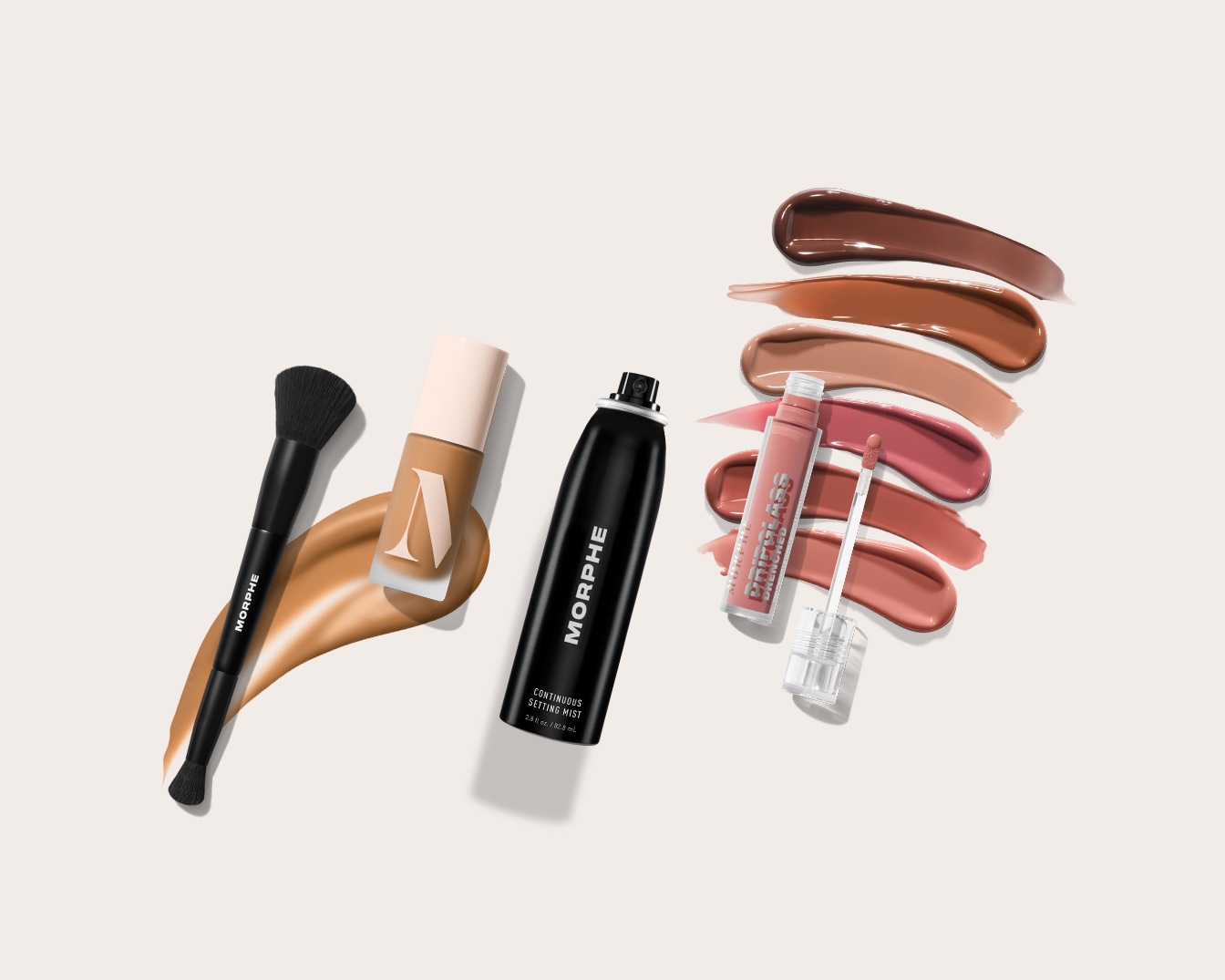 Na zdjęciu widać pędzel do makijażu Morphe, podkład w płynie, utrwalacz w sprayu oraz błyszczyk do ust z różnymi odcieniami, idealne do stworzenia kompletnego makijażu.