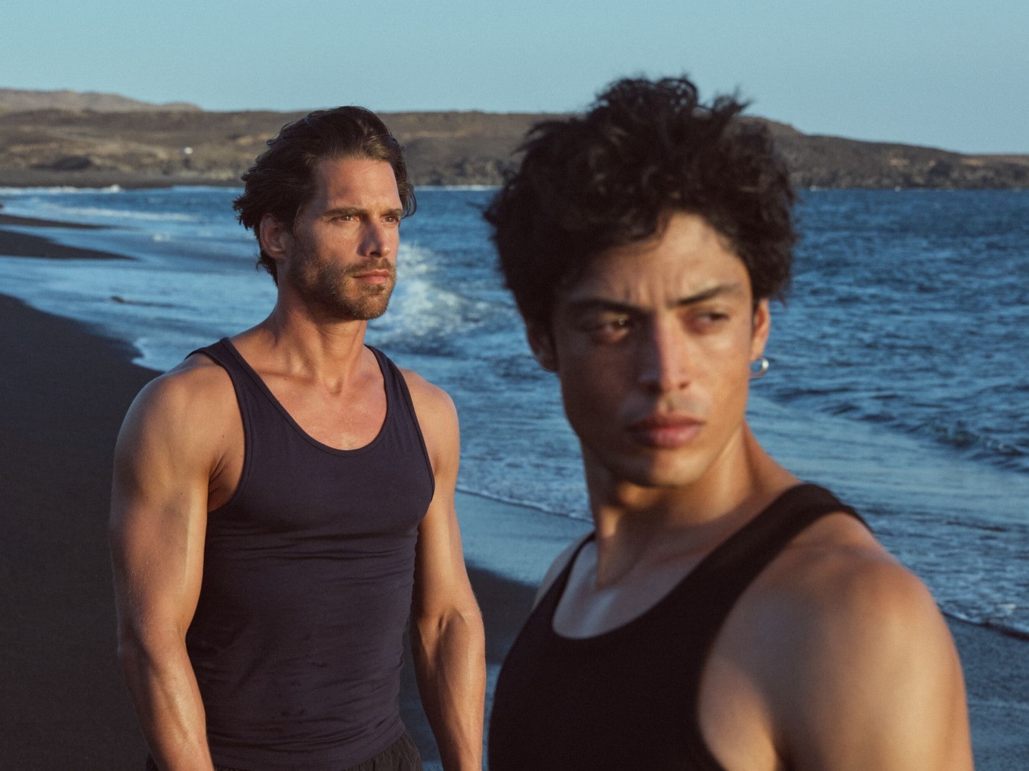 Zwei muskulöse Männer in Tanktops stehen am Strand mit dunklem Sand und blicken aufs Meer.