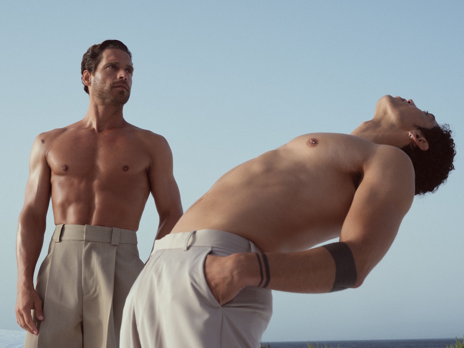 Zwei Männer mit freiem Oberkörper und beigen Hosen posieren vor einem hellblauen Himmel, wobei einer aufrecht steht und der andere sich nach hinten lehnt.