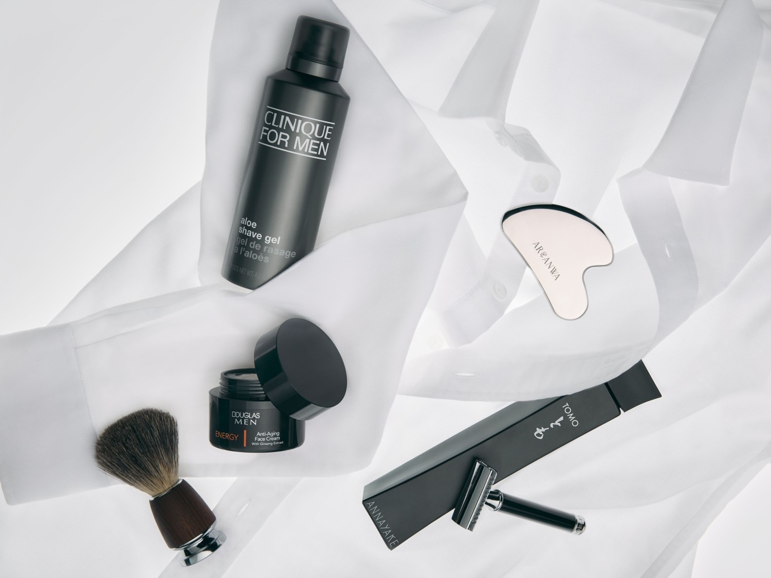 Auf einem weißen Hemd liegen verschiedene Herrenpflegeprodukte, darunter Clinique Aloe Shave Gel, Douglas Men Anti-Aging Face Cream, ein Rasierpinsel, ein Arganwa-Gesichtsmassagewerkzeug und ein Tomo-Rasierhobel von Annayake.