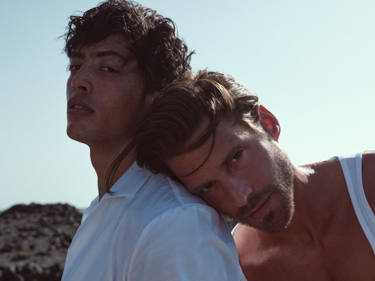 Zwei Männer posieren eng beieinander, einer mit weißem Hemd und der andere mit weißem Tanktop, vor einem hellblauen Himmel.