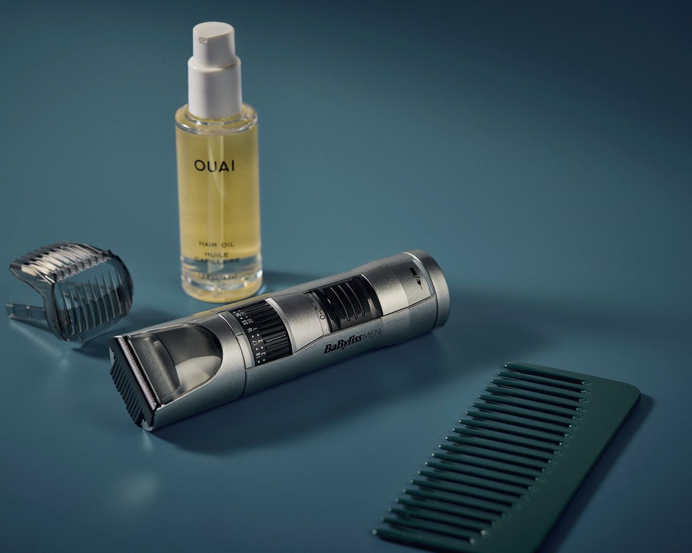 Auf blauem Grund liegen ein silberner Haarschneider von BabylissMEN, eine Flasche Haaröl von OUAI, ein Trimmeraufsatz und ein grüner Kamm.