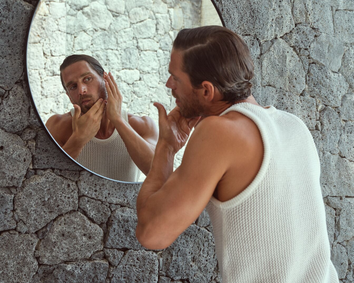Ein Mann im weißen Tanktop betrachtet sich im runden Spiegel und berührt sein Gesicht vor einer grauen Steinwand.