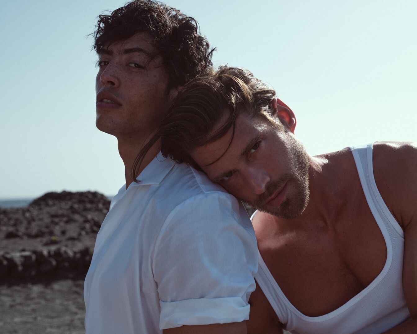 Zwei Männer posieren vor einem sonnigen Himmel, einer trägt ein weißes Hemd, der andere ein weißes Tanktop, beide mit einem entspannten und vertrauten Ausdruck.