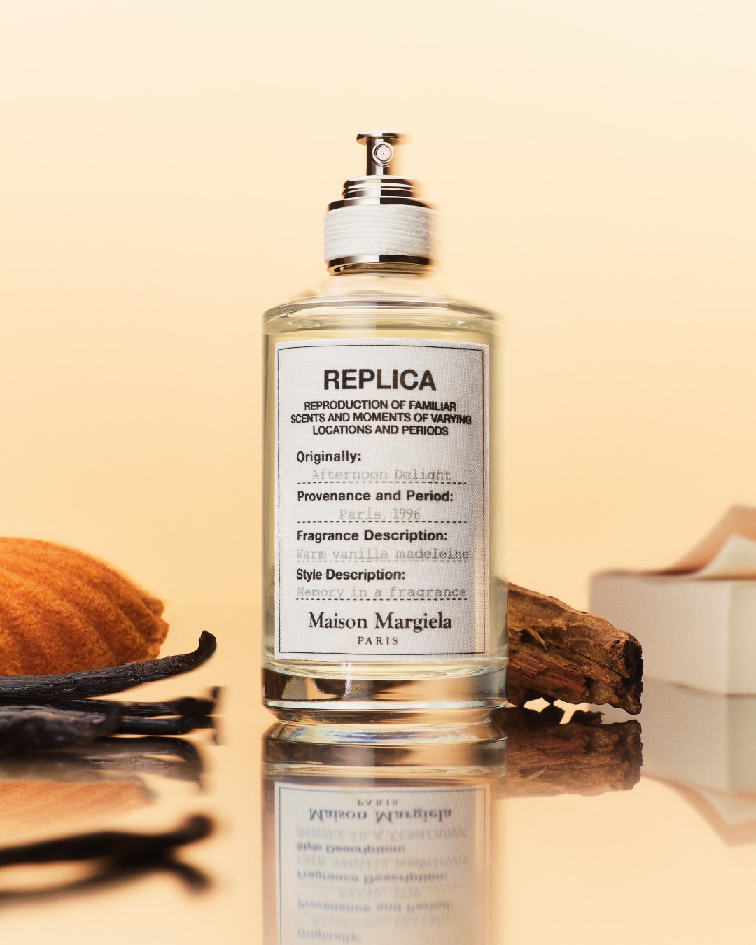 Przezroczysty flakon perfum Replica Maison Margiela Paris z etykietą opisującą zapach jako „Warm vanilla madeleine”, obok madlenki i laski wanilii.