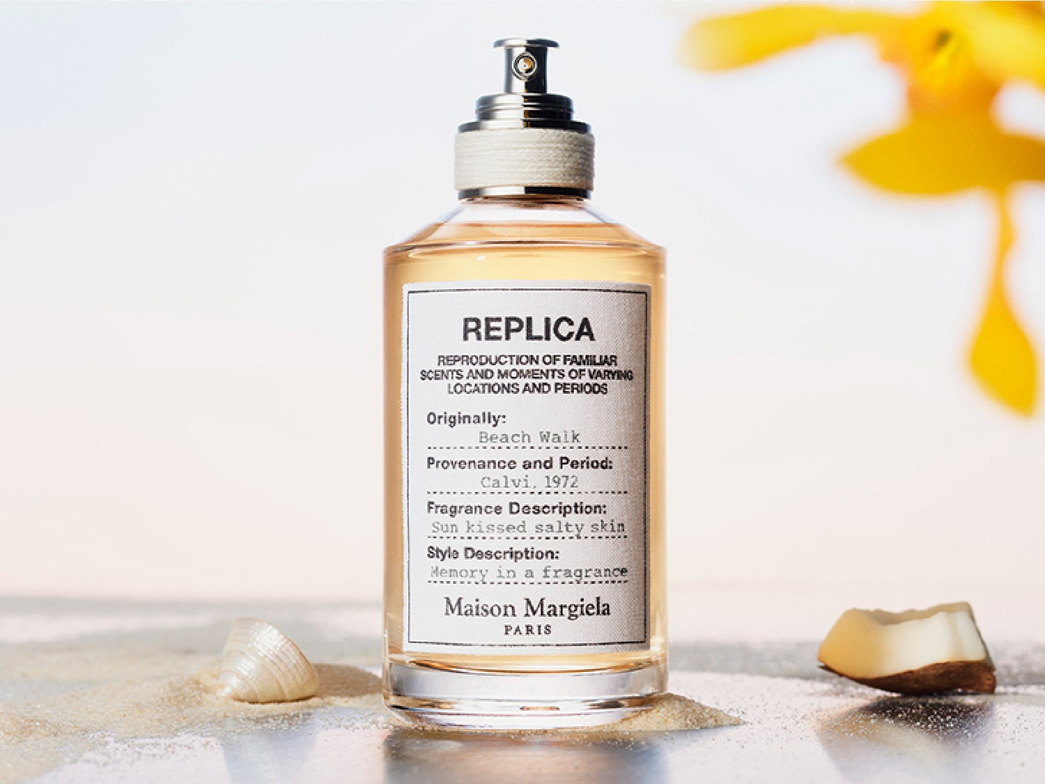 Butelka perfum Replica Beach Walk marki Maison Margiela, z etykietą opisującą zapach jako "sun kissed salty skin", stoi na piasku obok muszelki i kawałka kokosa.