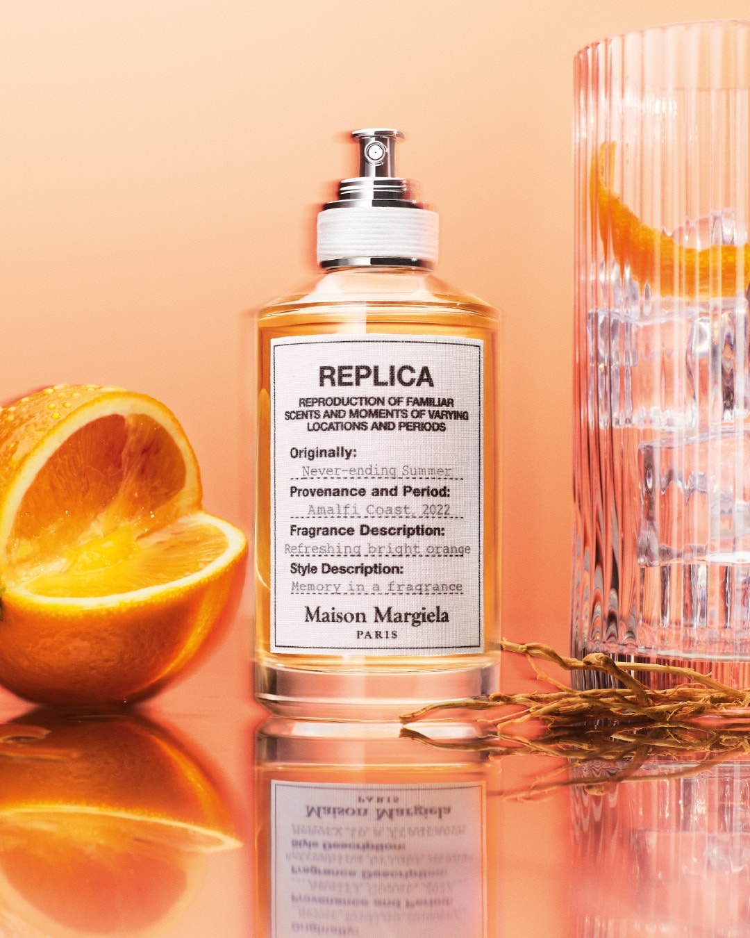 Butelka perfum Replica Maison Margiela Paris o zapachu Never-ending Summer, Amalfi Coast 2022, z nutami orzeźwiającej, jasnej pomarańczy, obok której leży połówka pomarańczy i szklanka z lodem i pomarańczą.