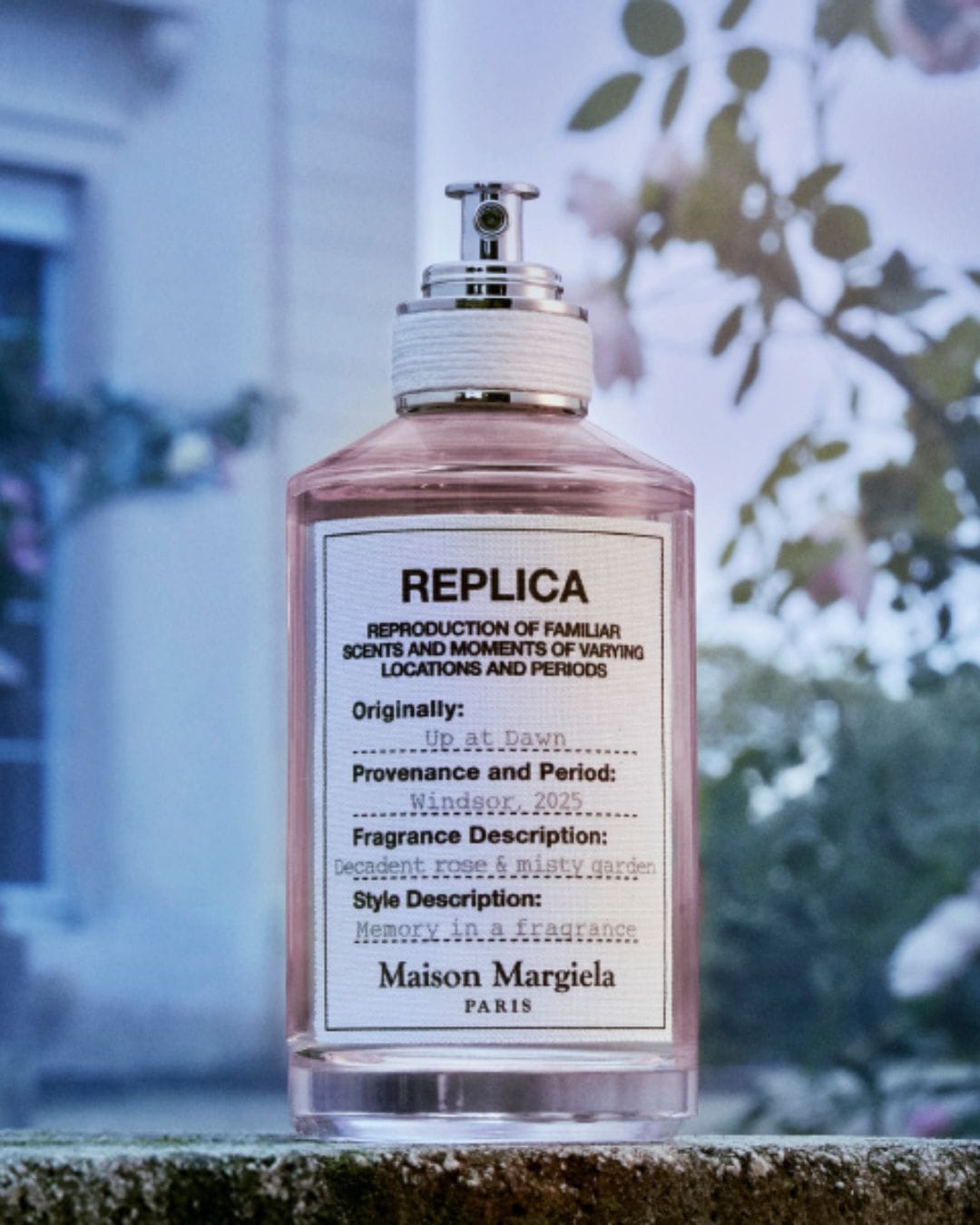 Butelka perfum Replica Maison Margiela Paris, o zapachu "Up at Dawn", z opisem "Decadent rose & misty garden", umieszczona na tle kwiatów.