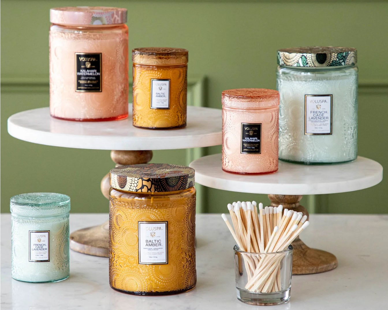 Różne świece Voluspa w ozdobnych słoikach, w tym zapachy takie jak Kalahari Watermelon, Baltic Amber i French Cade Lavender, ustawione na dwupoziomowym stojaku.