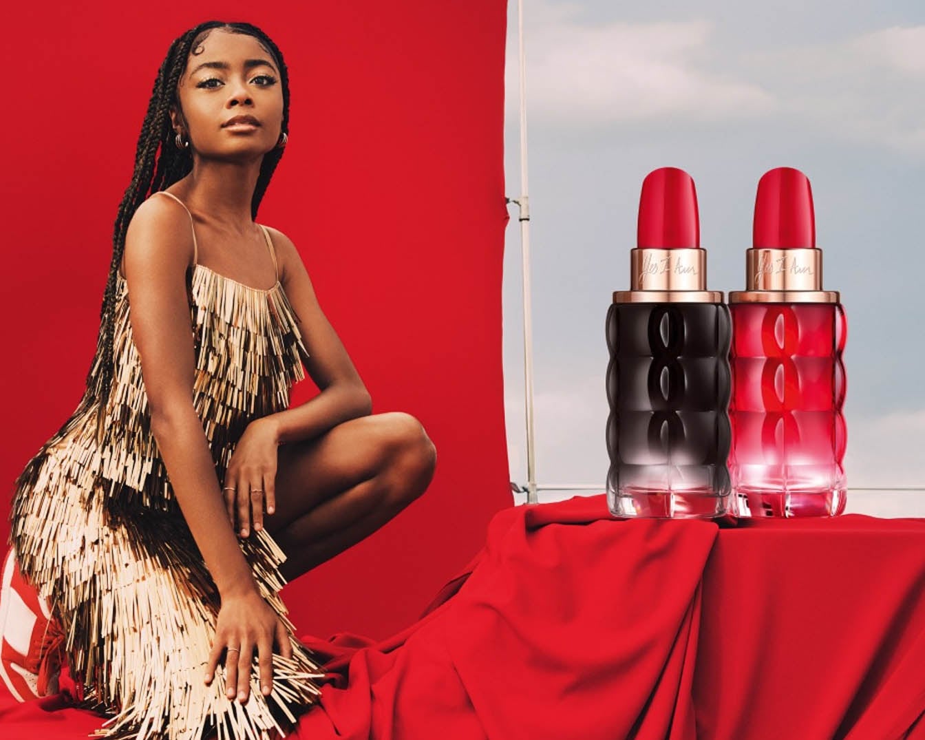 Na czerwonym tle kobieta w złotej sukience pozuje obok dwóch flakonów perfum Les Iles Kisses w odcieniach czerni i czerwieni, które kuszą swoim zapachem.