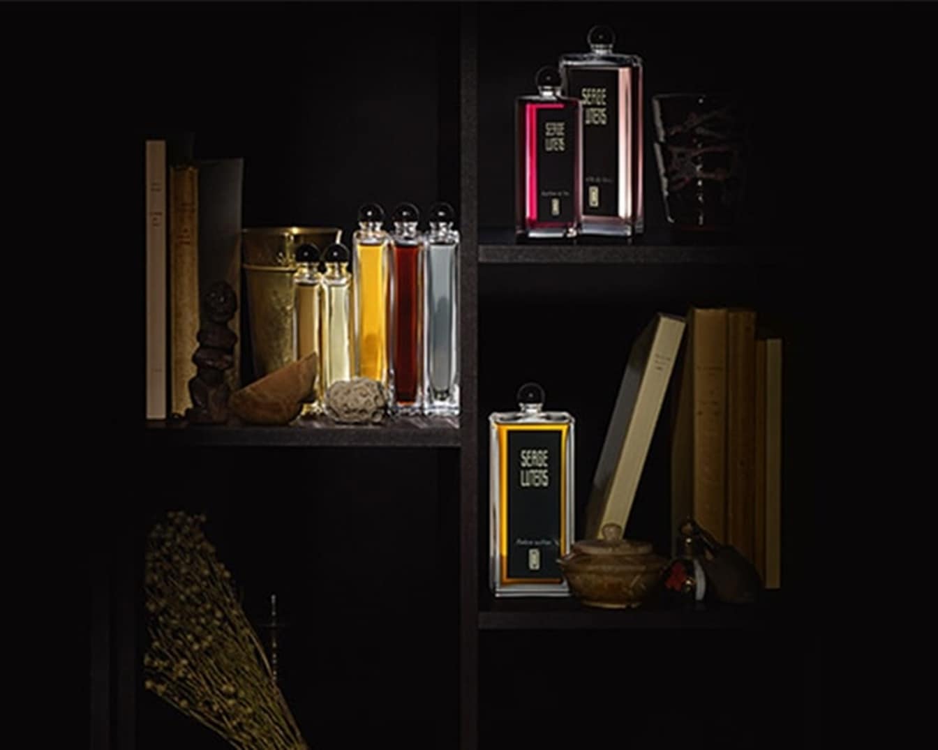 Auf einem dunklen Regal stehen verschiedene Flakons von Serge Lutens, einige mit goldenen, andere mit roten Akzenten, umgeben von Büchern und dekorativen Elementen.