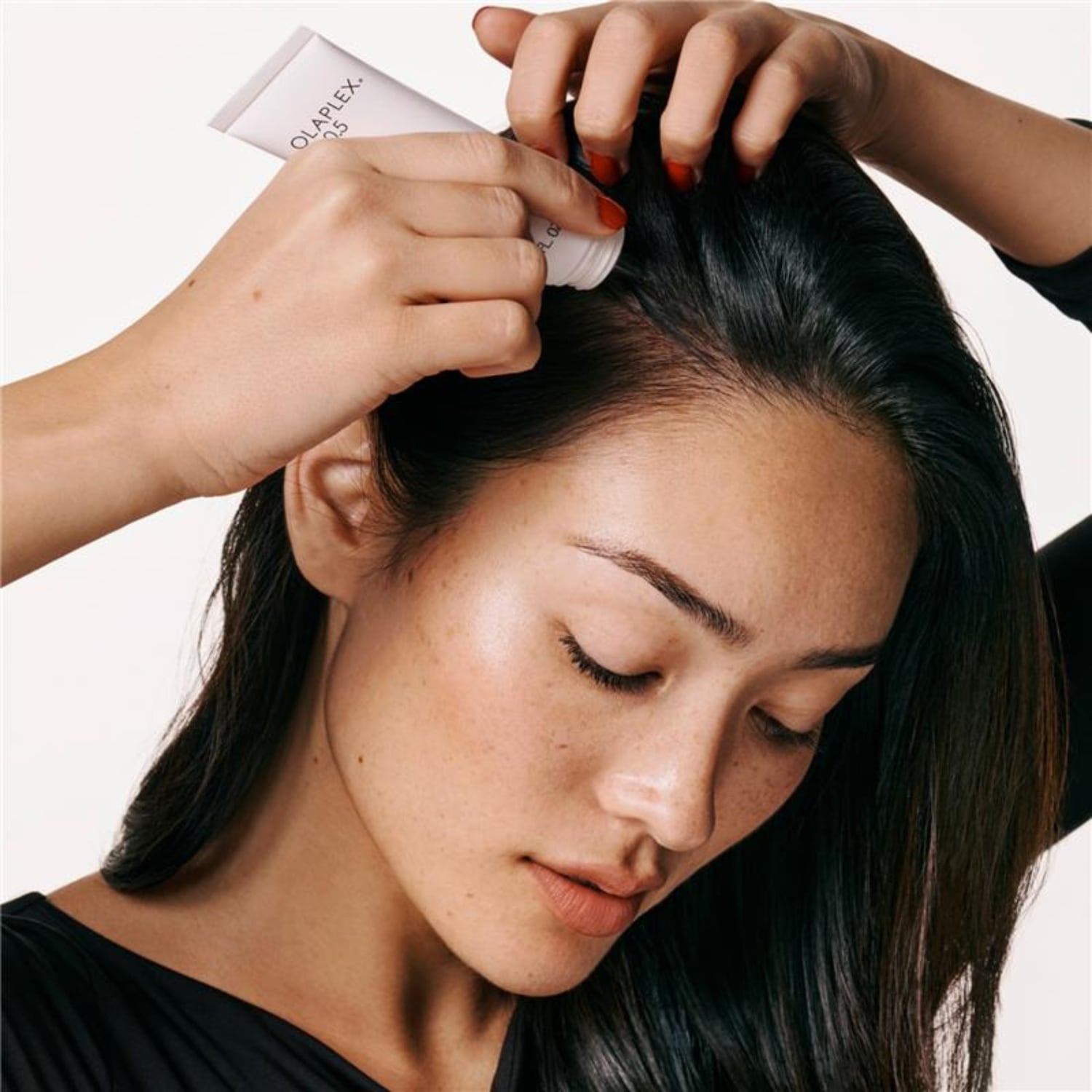 Kobieta z ciemnymi włosami aplikuje białą odżywkę Olaplex No. 0 na skórę głowy, trzymając tubkę w dłoniach z czerwonym manicure.