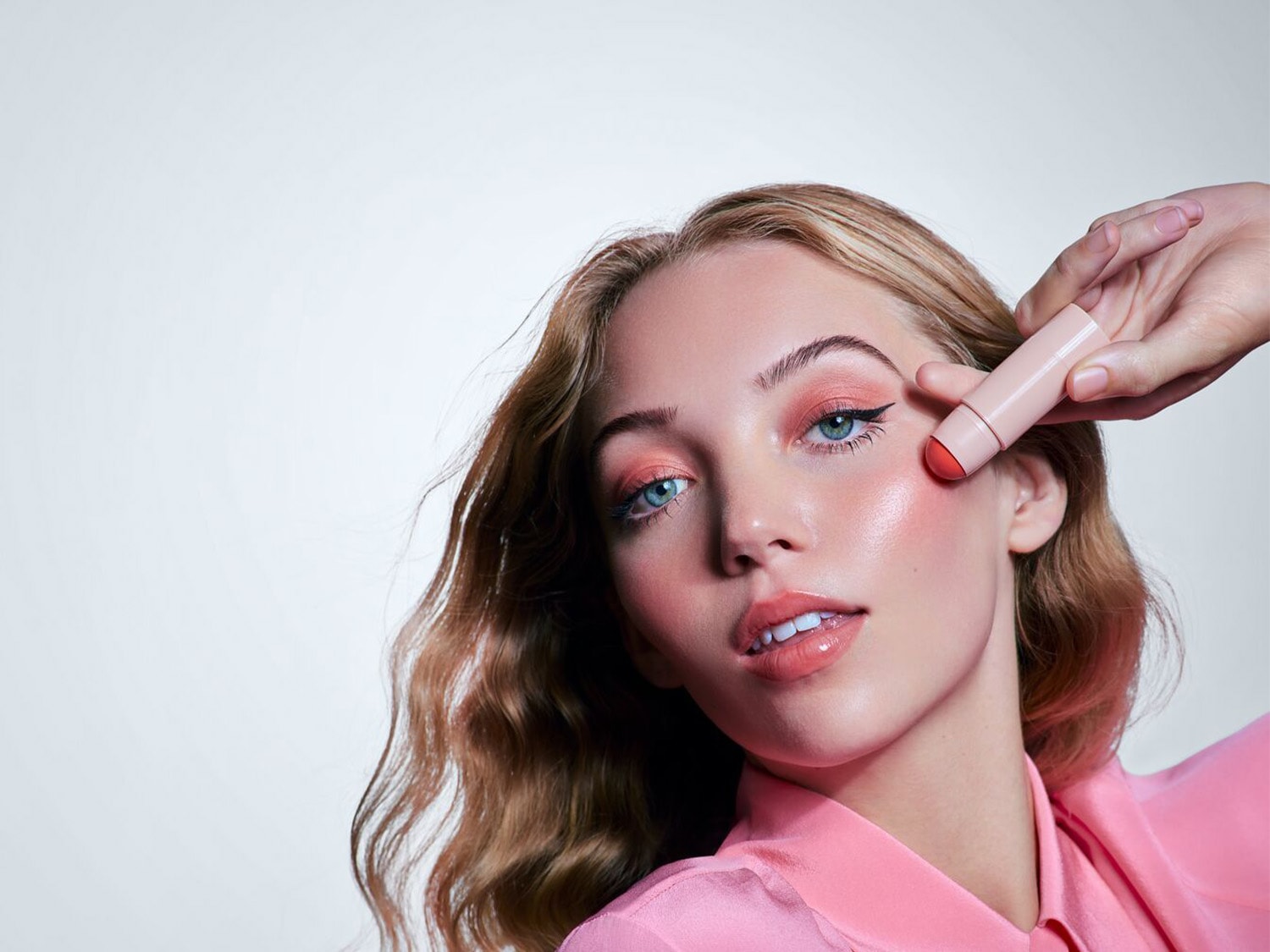 Een jonge vrouw met blond haar en blauwe ogen brengt een roze blushstick aan op haar wang voor een frisse en stralende look.