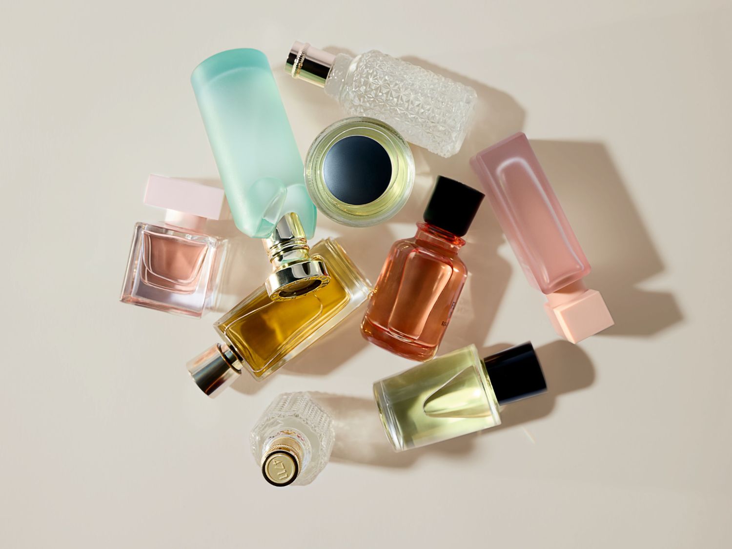 Een verzameling parfumflesjes in verschillende vormen en kleuren, waaronder roze, blauw, goud en amber, verspreid over een zachte, crèmekleurige achtergrond.