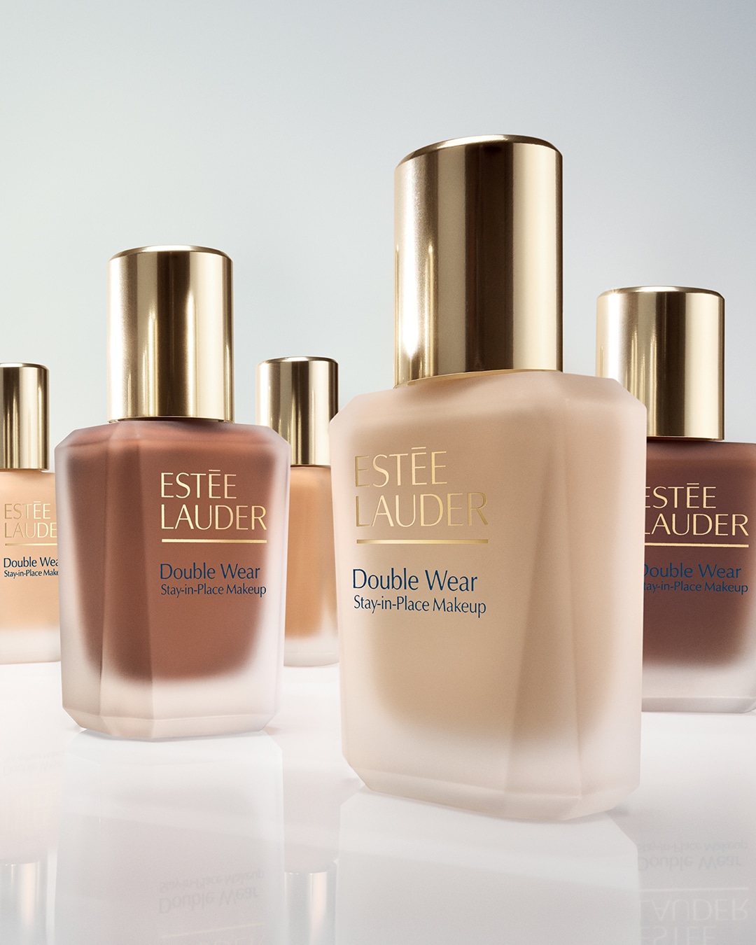Eleganckie flakony podkładu Estée Lauder Double Wear Stay-in-Place Makeup w różnych odcieniach, z matowego szkła i złotymi nakrętkami, prezentują się na błyszczącej powierzchni.