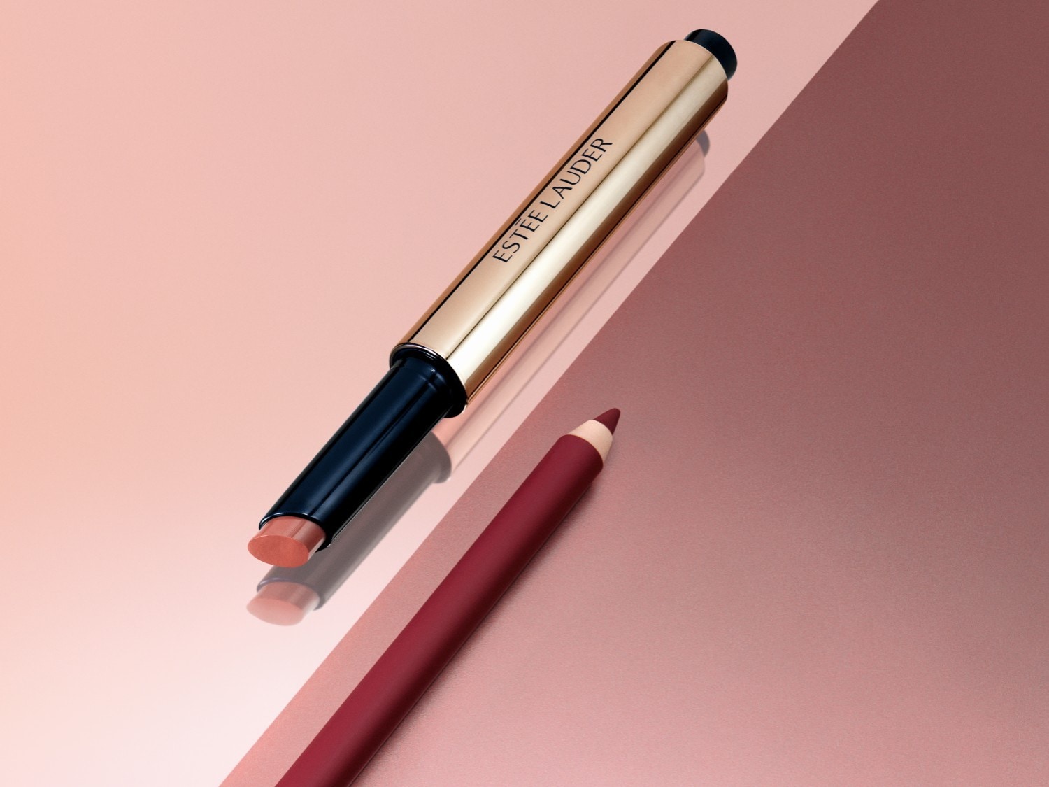 Szminka Estée Lauder w złotym opakowaniu leży obok konturówki do ust w odcieniu czerwonego wina na tle w kolorze różowego złota.