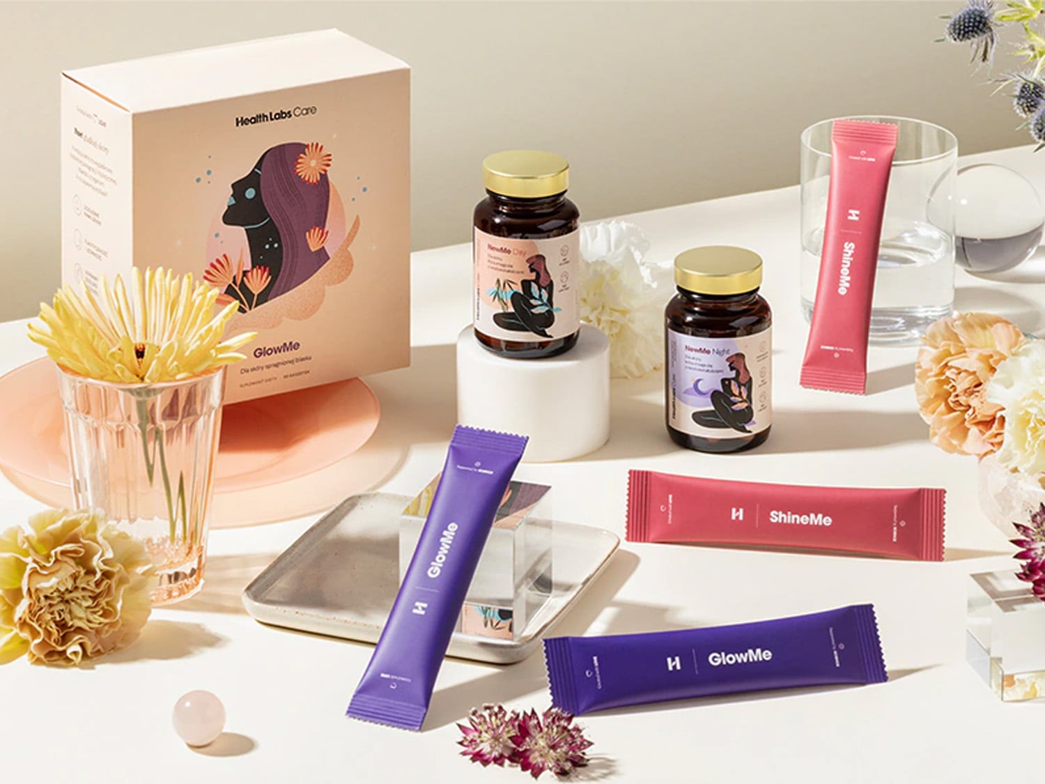 Na białym tle znajdują się suplementy Health Labs Care, w tym GlowMe w fioletowym opakowaniu i ShineMe w różowym, obok nich stoją flakony NewMe Day i NewMe Night.