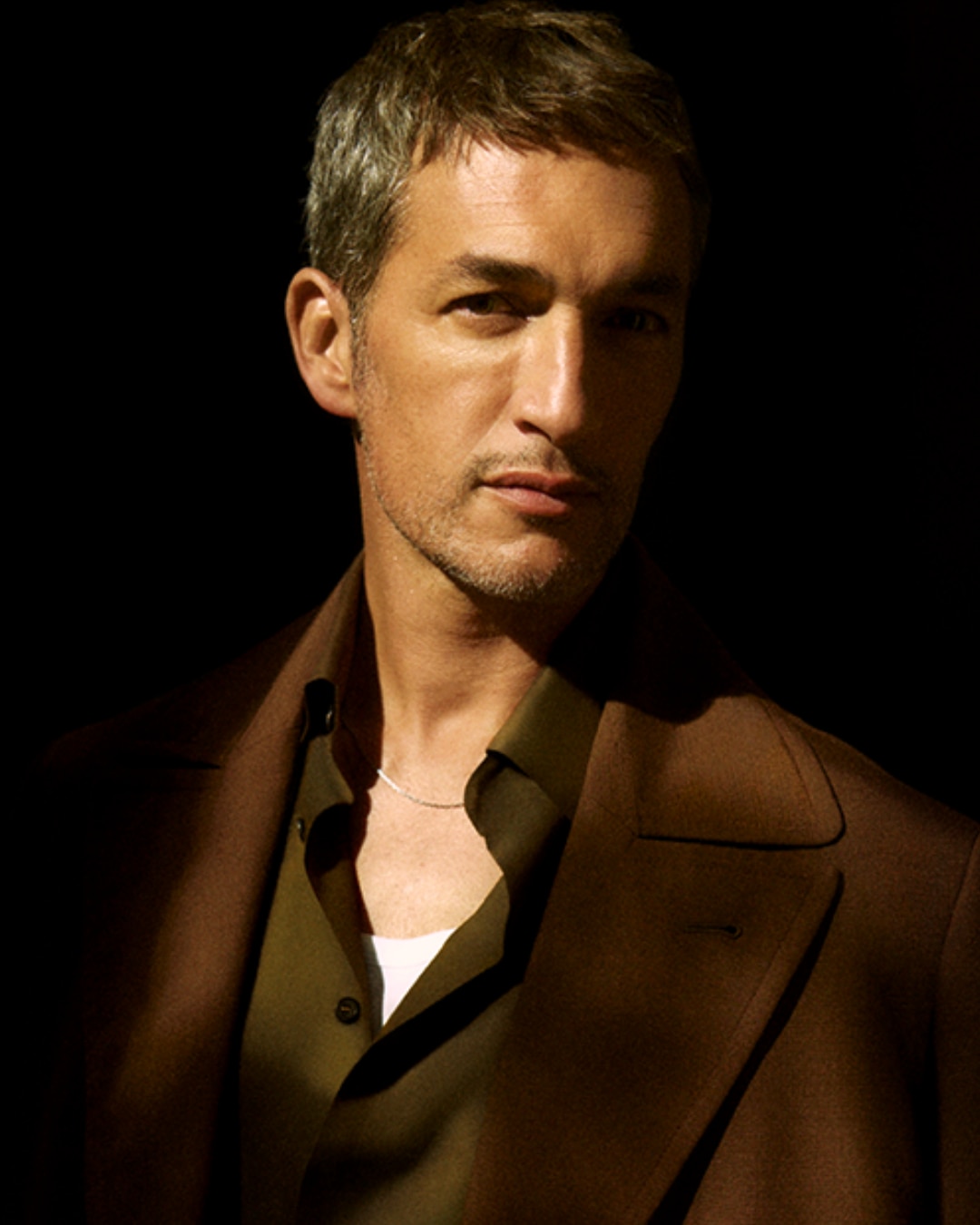 Portret mężczyzny w brązowym płaszczu i zielonej koszuli, z srebrnym łańcuszkiem na szyi, na ciemnym tle.