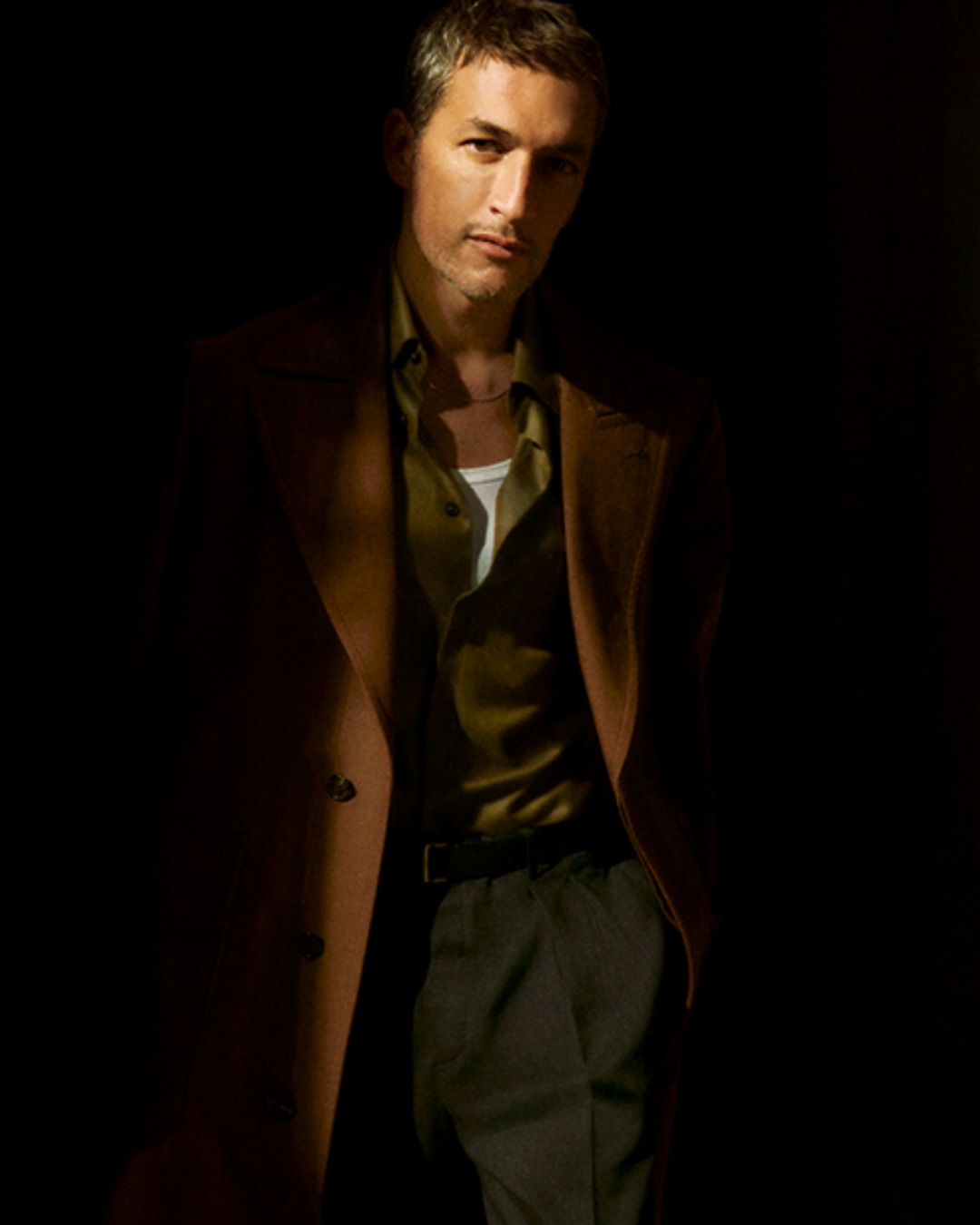 Mężczyzna w brązowym płaszczu i oliwkowej koszuli, z nonszalancko zarzuconym płaszczem na ramieniu, emanuje pewnością siebie.