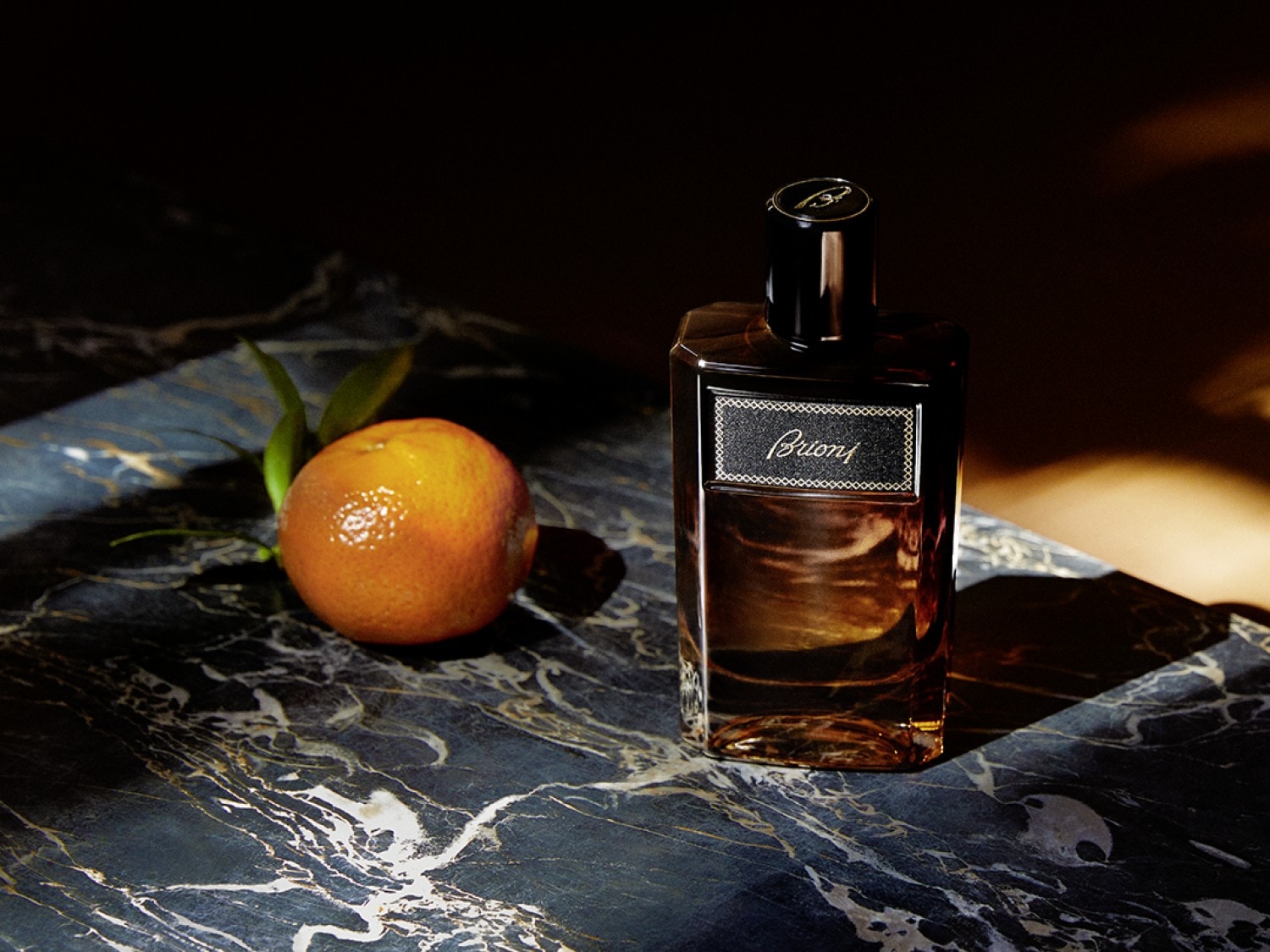 Na marmurowym tle stoi flakon perfum Brioni obok soczystej pomarańczy z zielonymi listkami, tworząc elegancką kompozycję.