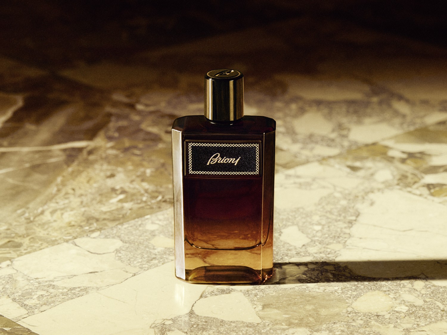 Elegancki flakon perfum Brioni o kwadratowym kształcie, z ciemnobrązowego szkła przechodzącego w bursztynowy odcień, z czarną etykietą i złotymi napisami, stoi na marmurowej powierzchni.