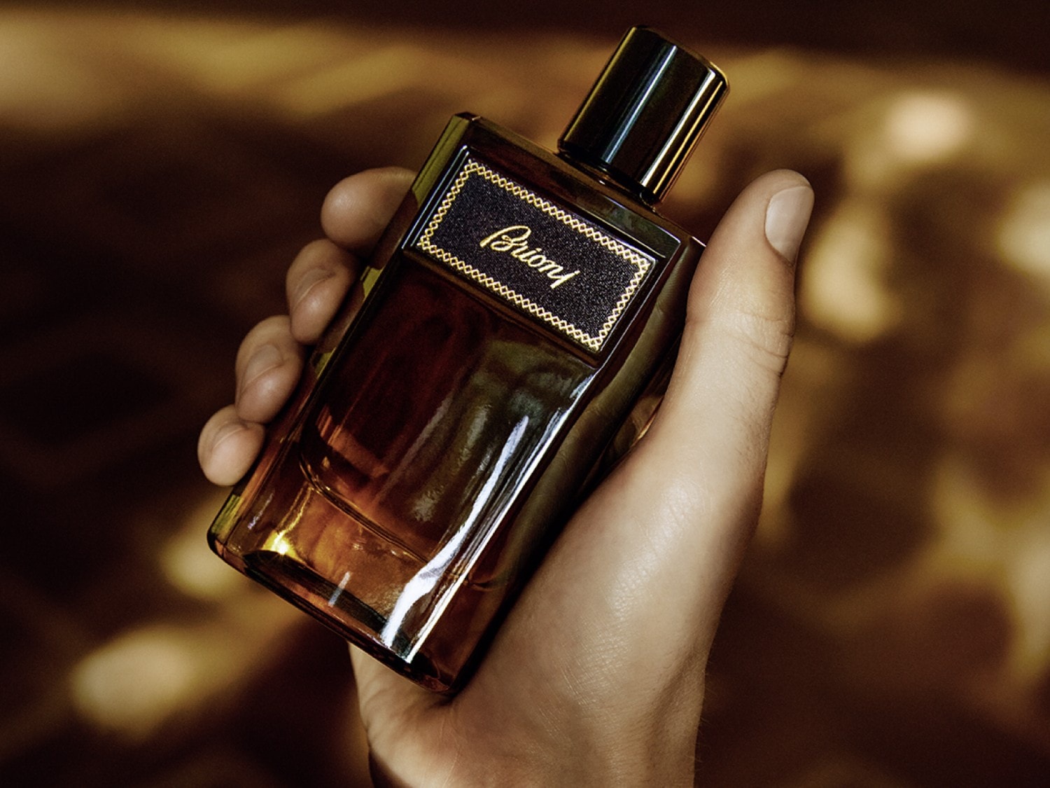 Na dłoni trzymany jest flakon perfum Brioni o bursztynowym odcieniu, z czarną etykietą i złotym napisem, emanujący luksusem i elegancją.