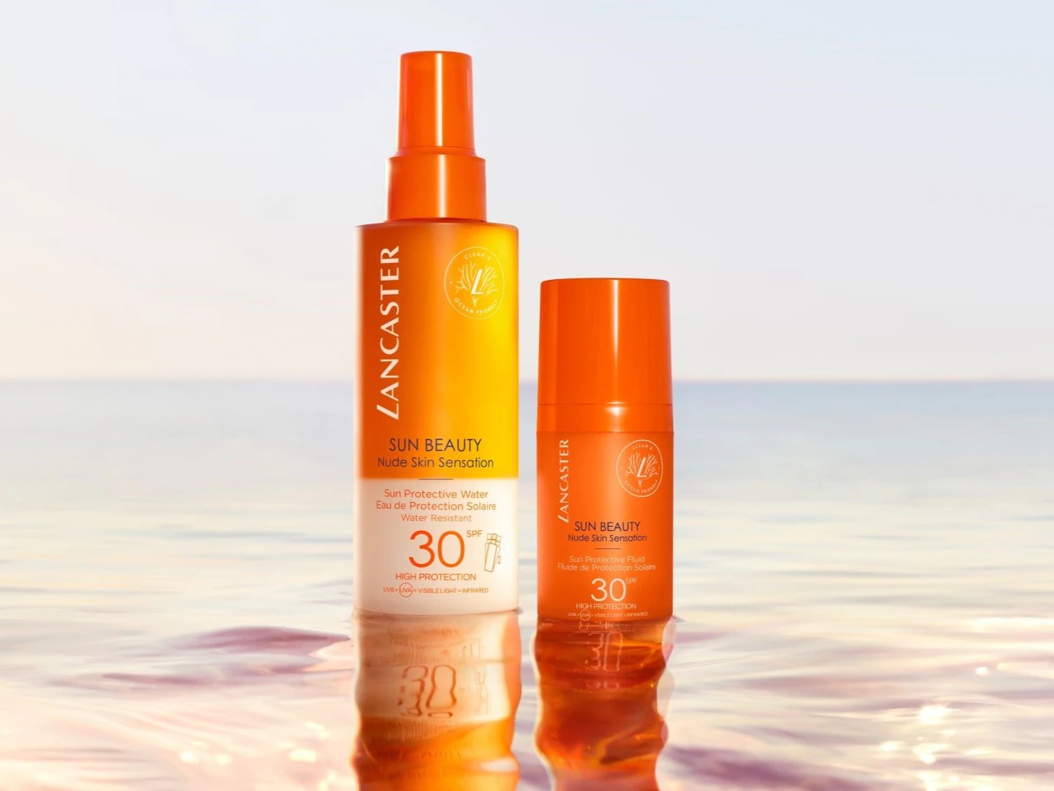 Twee oranje Lancaster Sun Beauty producten, een water en een fluid, beide met SPF 30, staan op een reflecterend oppervlak met de zee op de achtergrond.