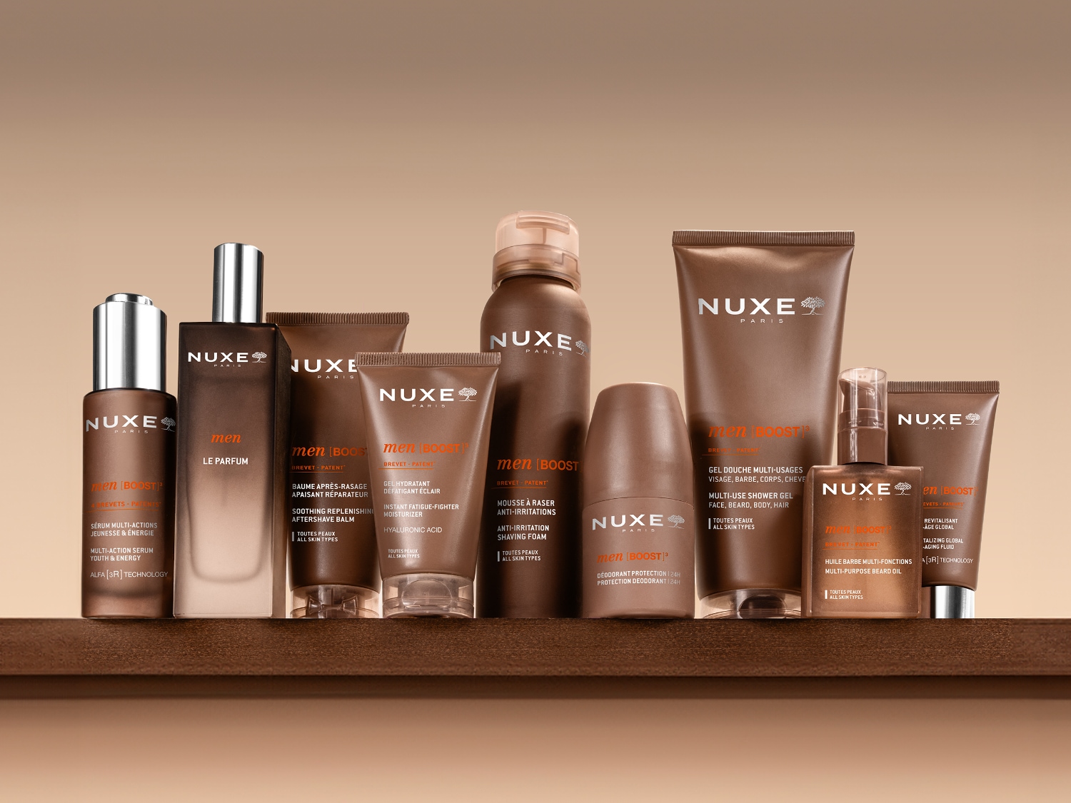 Braune Nuxe Men-Pflegeprodukte, darunter ein Multi-Use-Duschgel, ein Anti-Age-Fluid, ein 24-Stunden-Deodorant und ein feuchtigkeitsspendendes Mehrzweckgel, liegen auf einem weißen Hemd mit einer schwarzen Fliege.