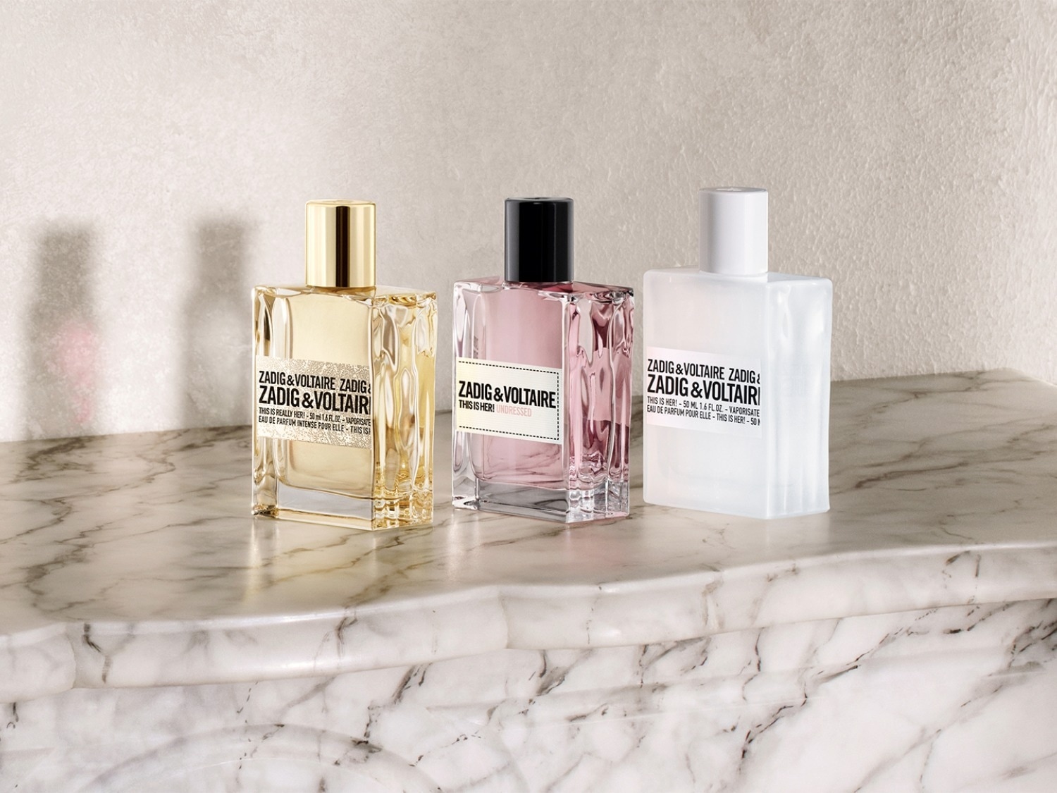Drie Zadig & Voltaire parfumflessen staan op een marmeren oppervlak, elk met een unieke kleur: goud, roze en wit.