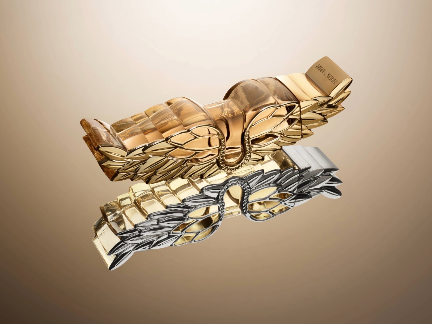 Gouden Zadig & Voltaire parfumfles in de vorm van een vleugel, liggend op een spiegelend oppervlak dat een zilveren reflectie creëert.