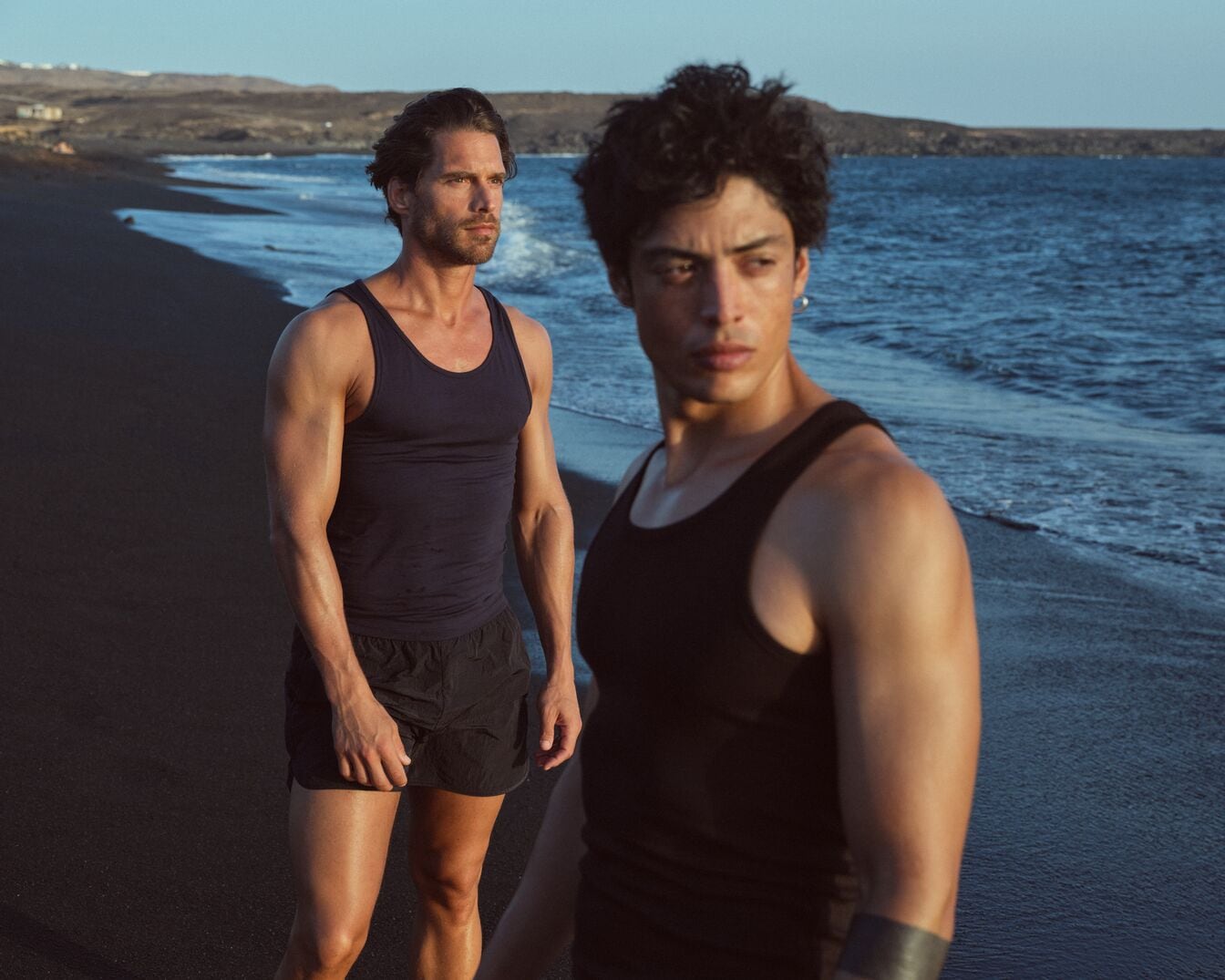 Zwei muskulöse Männer in schwarzen Tanktops und Shorts stehen am Strand mit dunklem Sand und blauem Meer.