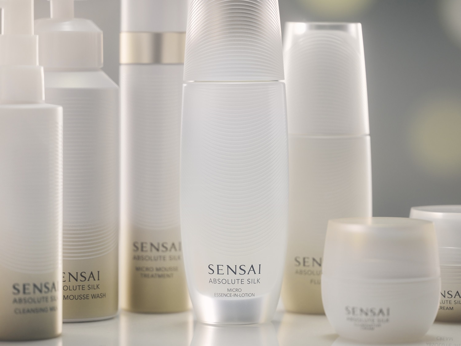 Różne produkty Sensai Absolute Silk w białych butelkach, w tym płyn do demakijażu, pianka do mycia, mus do zabiegów, esencja w lotionie, fluid i krem.