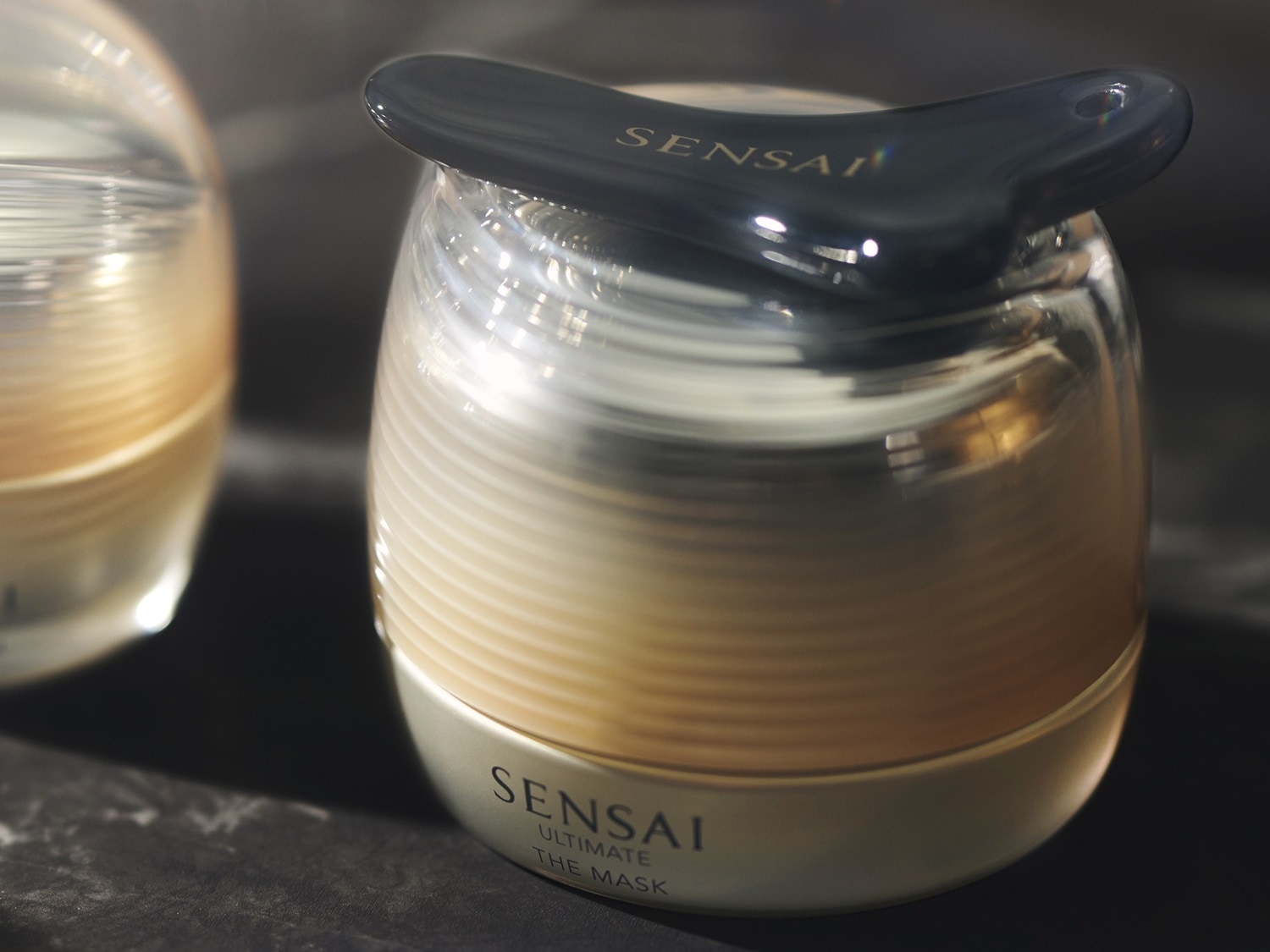 Słoik z maską Sensai Ultimate The Mask w złotym opakowaniu i czarnym aplikatorem na górze.