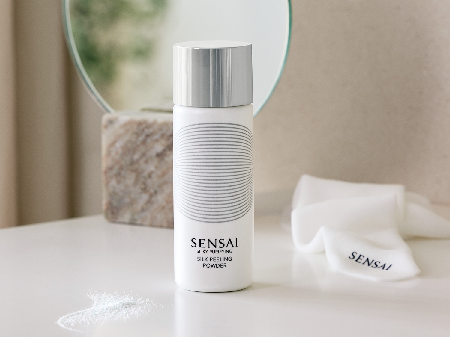 Biała butelka Sensai Silky Purifying Silk Peeling Powder z srebrną nakrętką, obok leży biała ściereczka z logo Sensai, a na blacie rozsypany jest biały proszek.