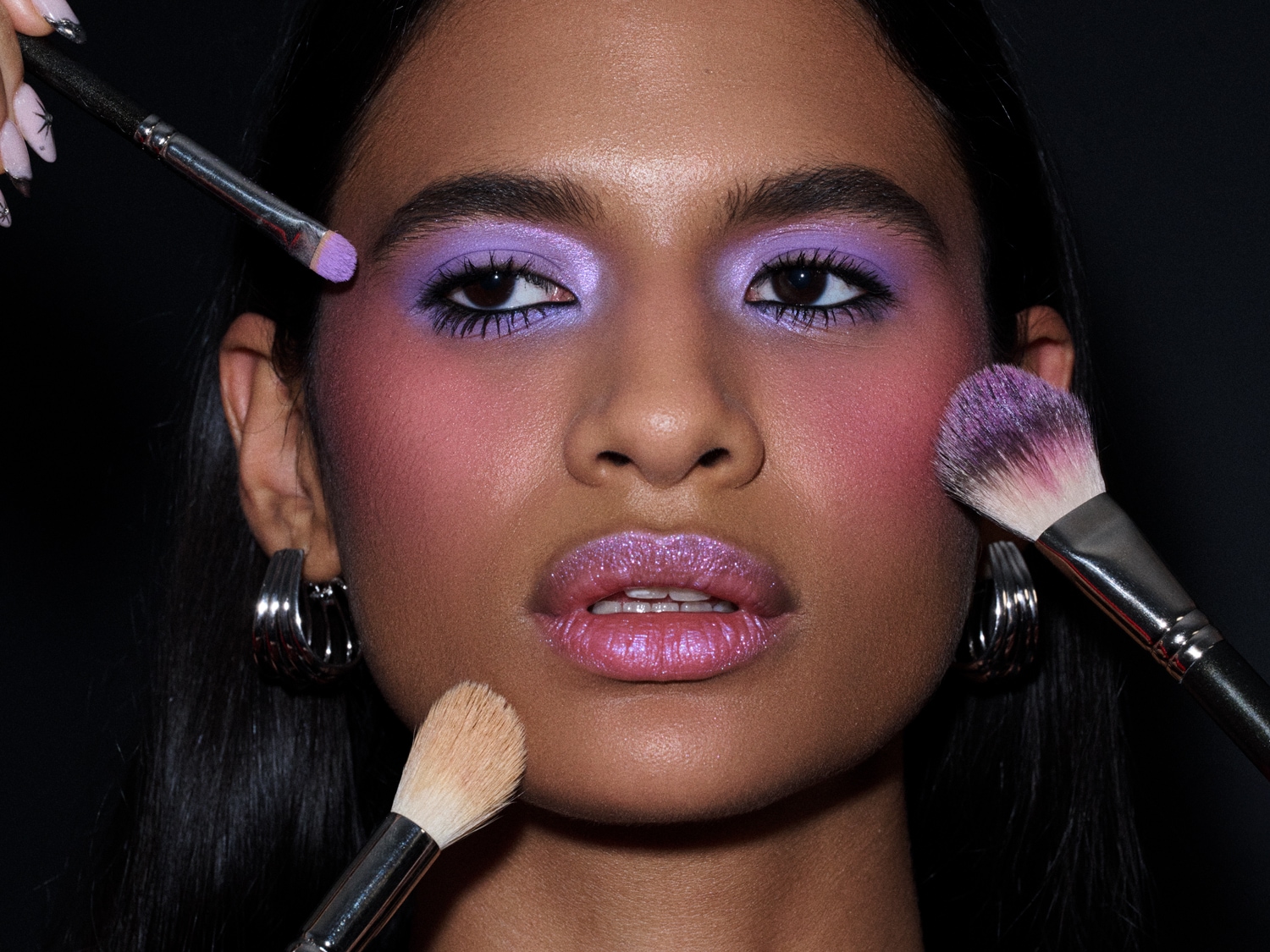 Een close-up van een vrouw met paarse oogschaduw, roze blush en glanzende lippen, terwijl make-upkwasten haar gezicht aanraken voor een complete schoonheidslook.