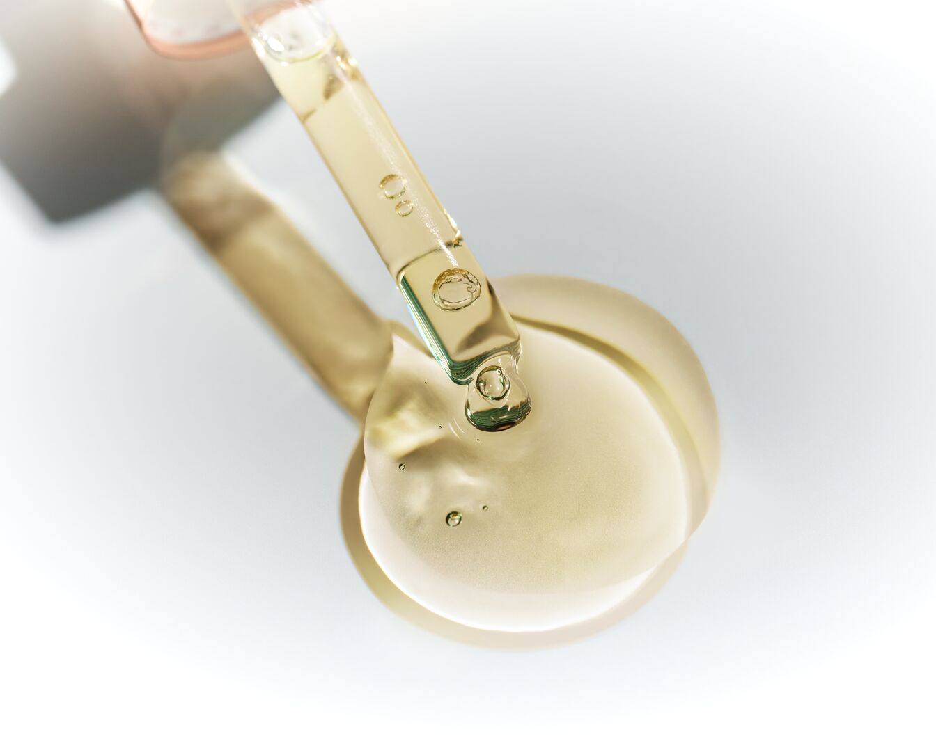 Ein durchsichtiger Glaspipette gibt einen Tropfen goldenes Serum auf eine glatte, hellbeige Oberfläche ab.