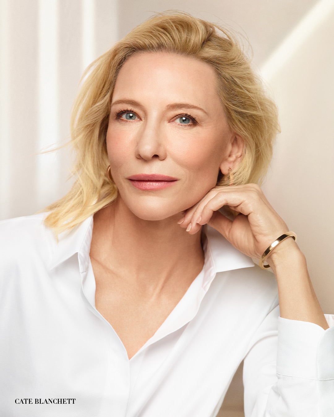 Cate Blanchett, z blond włosami i niebieskimi oczami, nosi białą koszulę i złotą bransoletkę, opierając podbródek na dłoni.