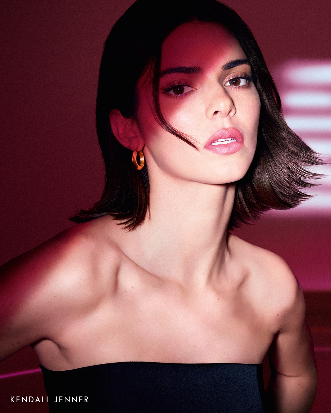 Portret Kendall Jenner z krótkimi, ciemnymi włosami i złotymi kolczykami na tle w odcieniach różu.