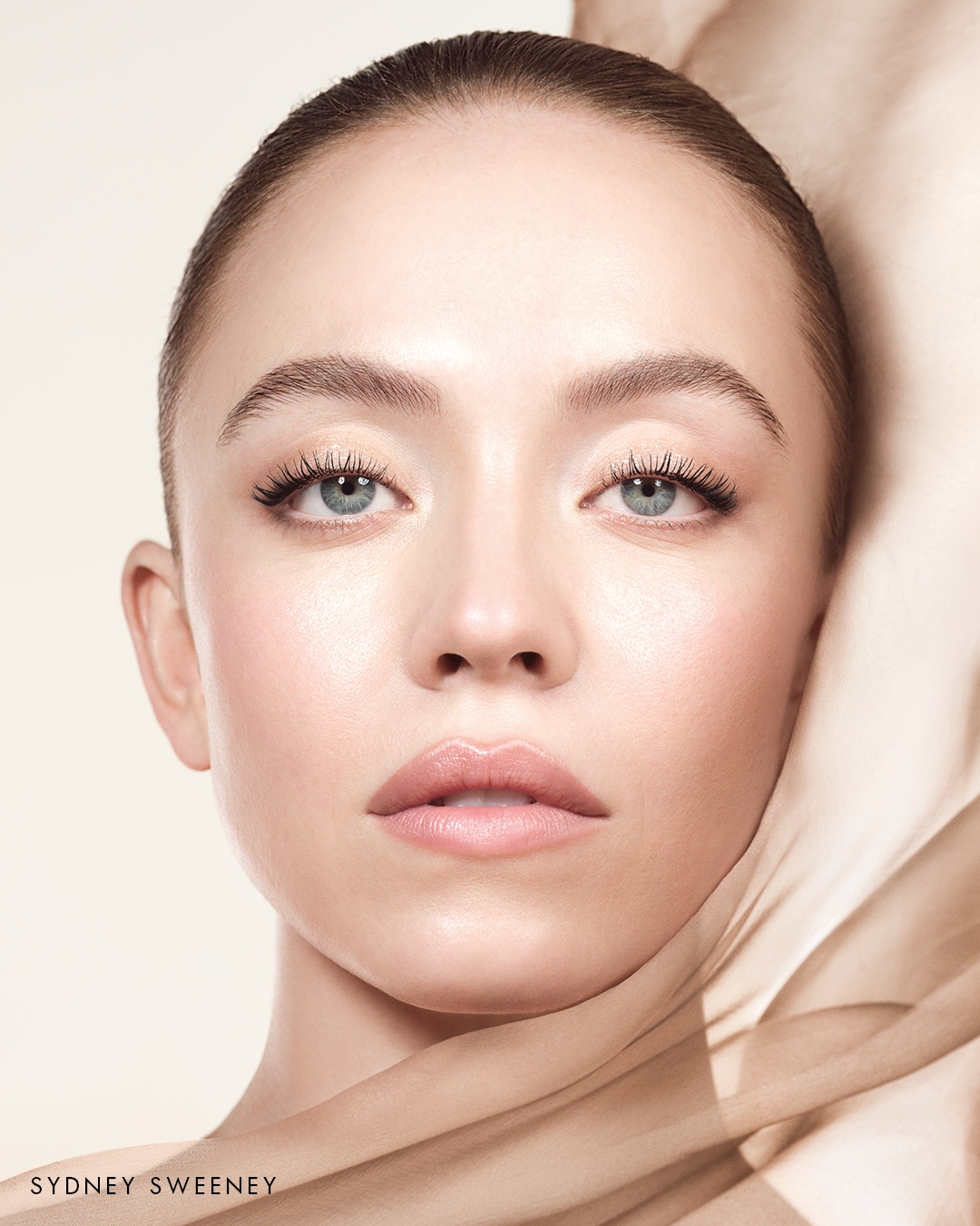 Portret kobiety o jasnej cerze, z niebieskimi oczami i naturalnym makijażem, otulonej beżową tkaniną.