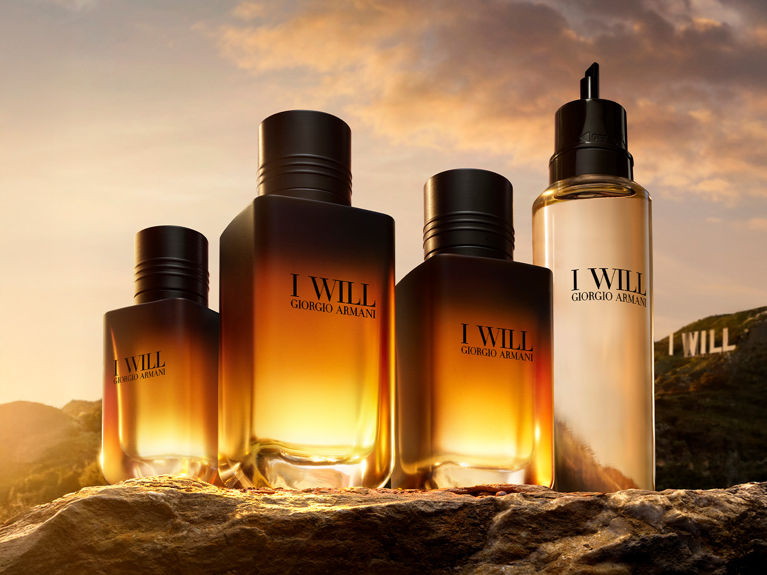 Cztery butelki perfum Giorgio Armani "I Will" w różnych rozmiarach, z czego trzy są prostokątne, a jedna cylindryczna, wszystkie z czarnymi nakrętkami, stoją na skalistej powierzchni na tle zachodzącego słońca i gór.