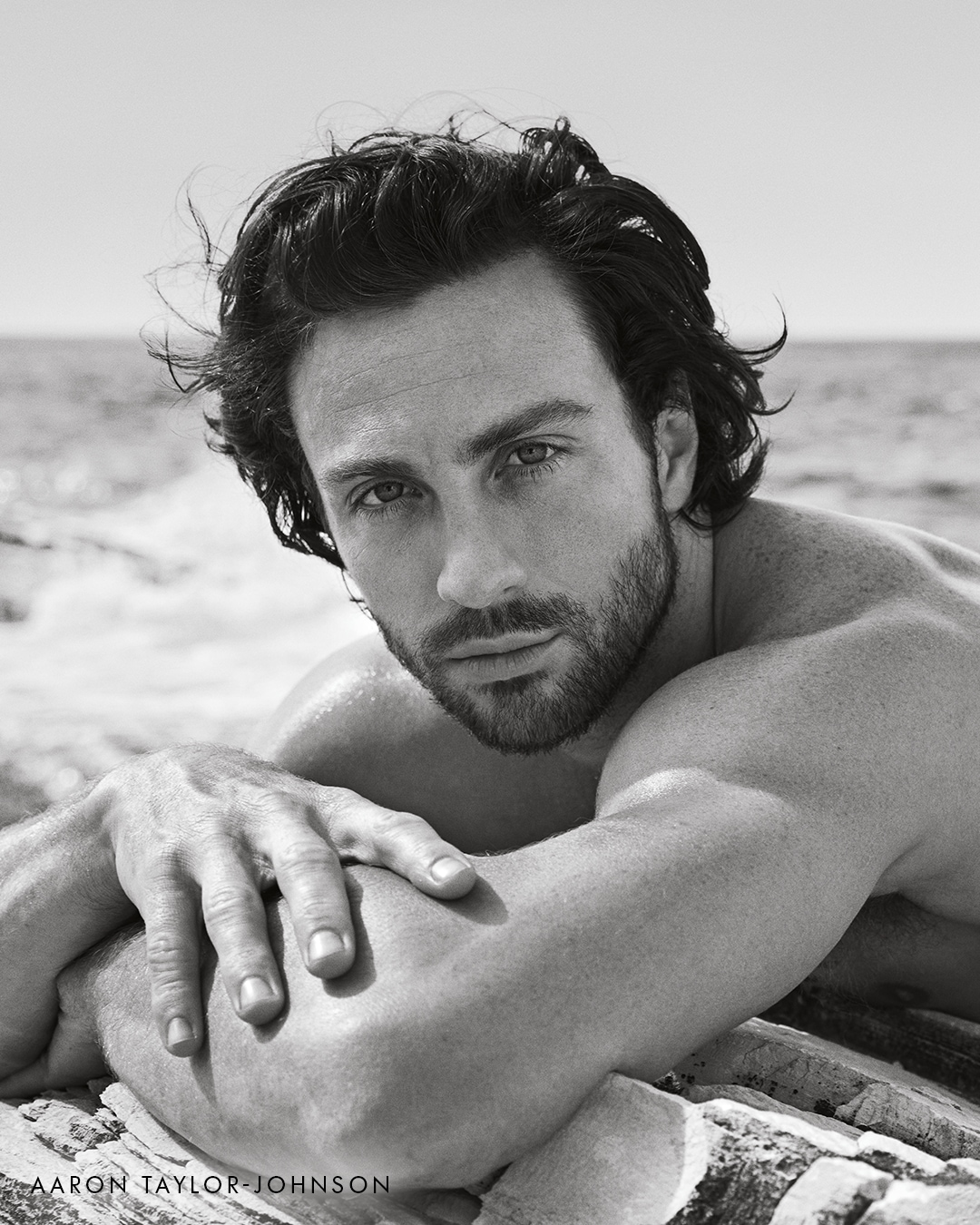 Czarno-białe zdjęcie Aarona Taylora-Johnsona leżącego na kamieniach z zamyślonym spojrzeniem.