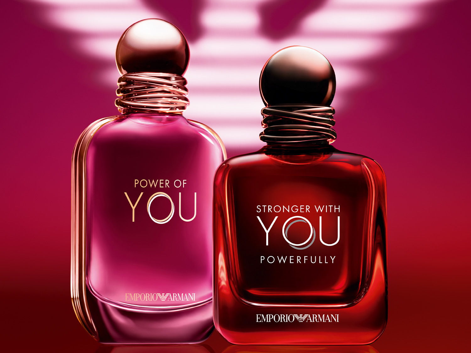 Dwa flakony perfum Emporio Armani, jeden w kolorze fuksji z napisem "POWER OF YOU", a drugi w kolorze czerwonym z napisem "STRONGER WITH YOU POWERFULLY", stojące na tle w odcieniach różu i czerwieni.