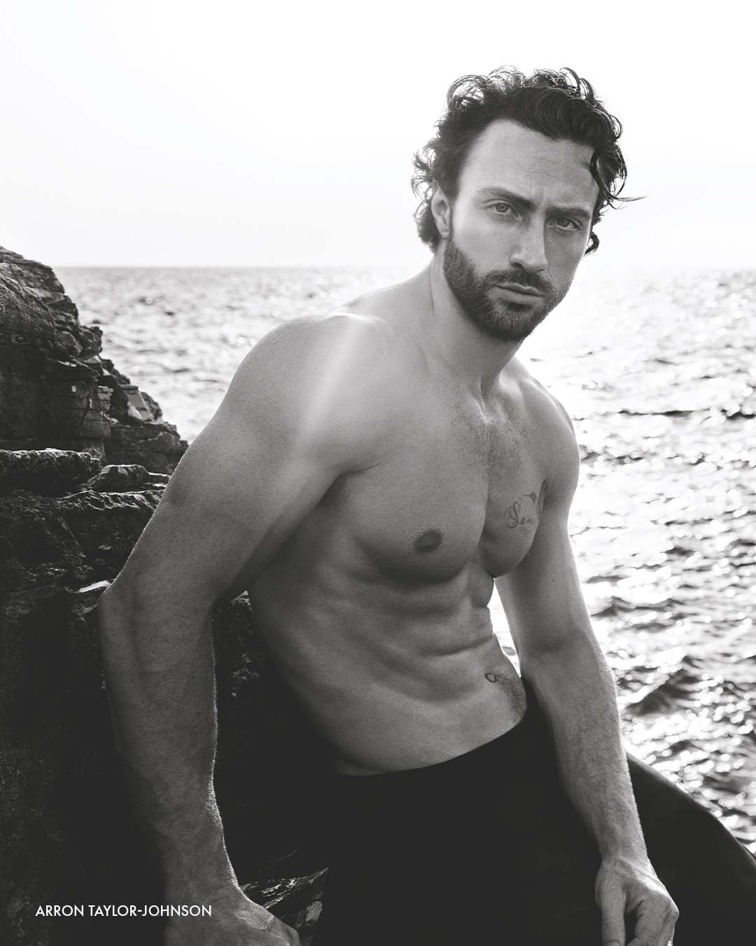Aaron Taylor-Johnson, z kręconymi włosami i brodą, pozuje bez koszuli na tle morza i skał, prezentując tatuaże na klatce piersiowej i brzuchu.