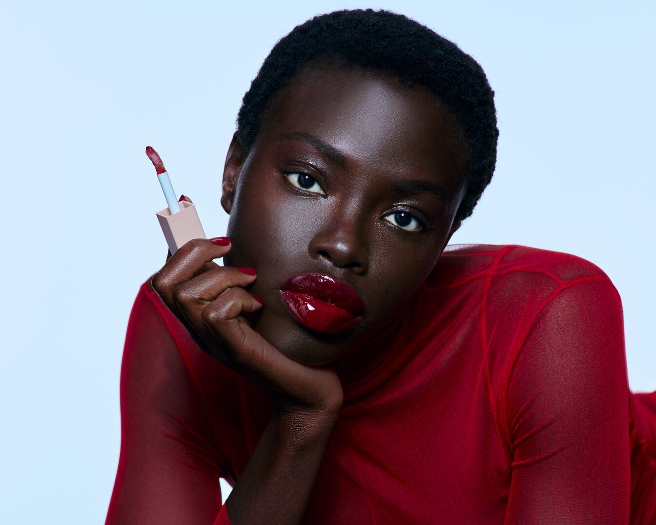 Eine Frau mit dunkler Haut und roten Lippen hält einen hellrosa Lipgloss mit einem hellblauen Applikator in der Hand.