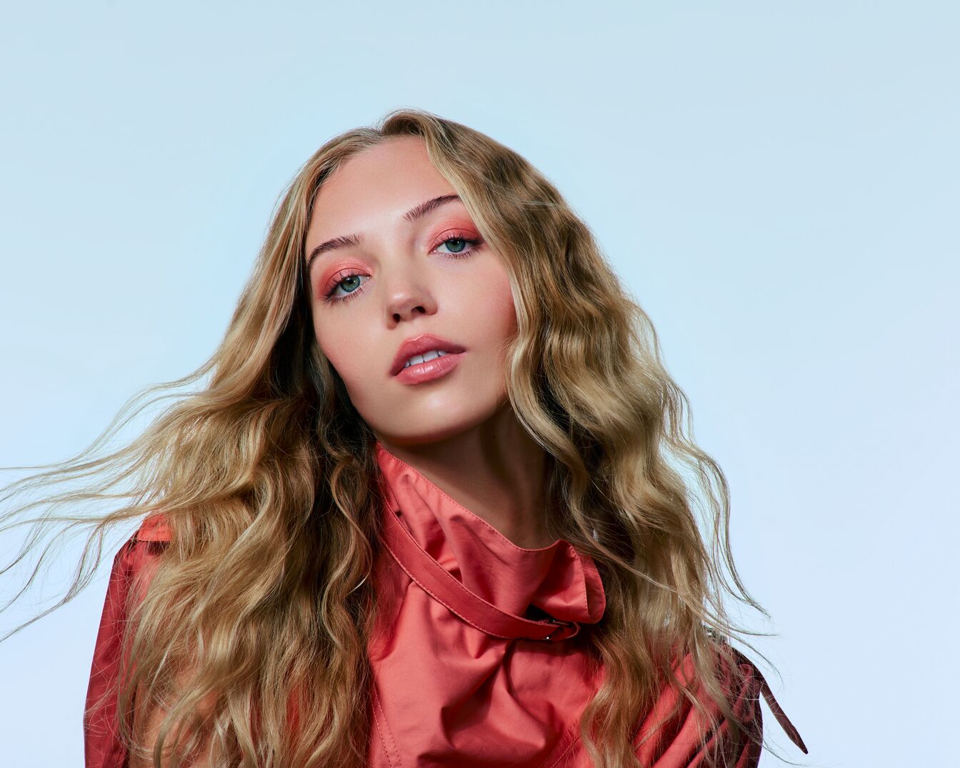 Une femme aux longs cheveux ondulés blond miel et au maquillage pêche pose avec une veste rose corail.