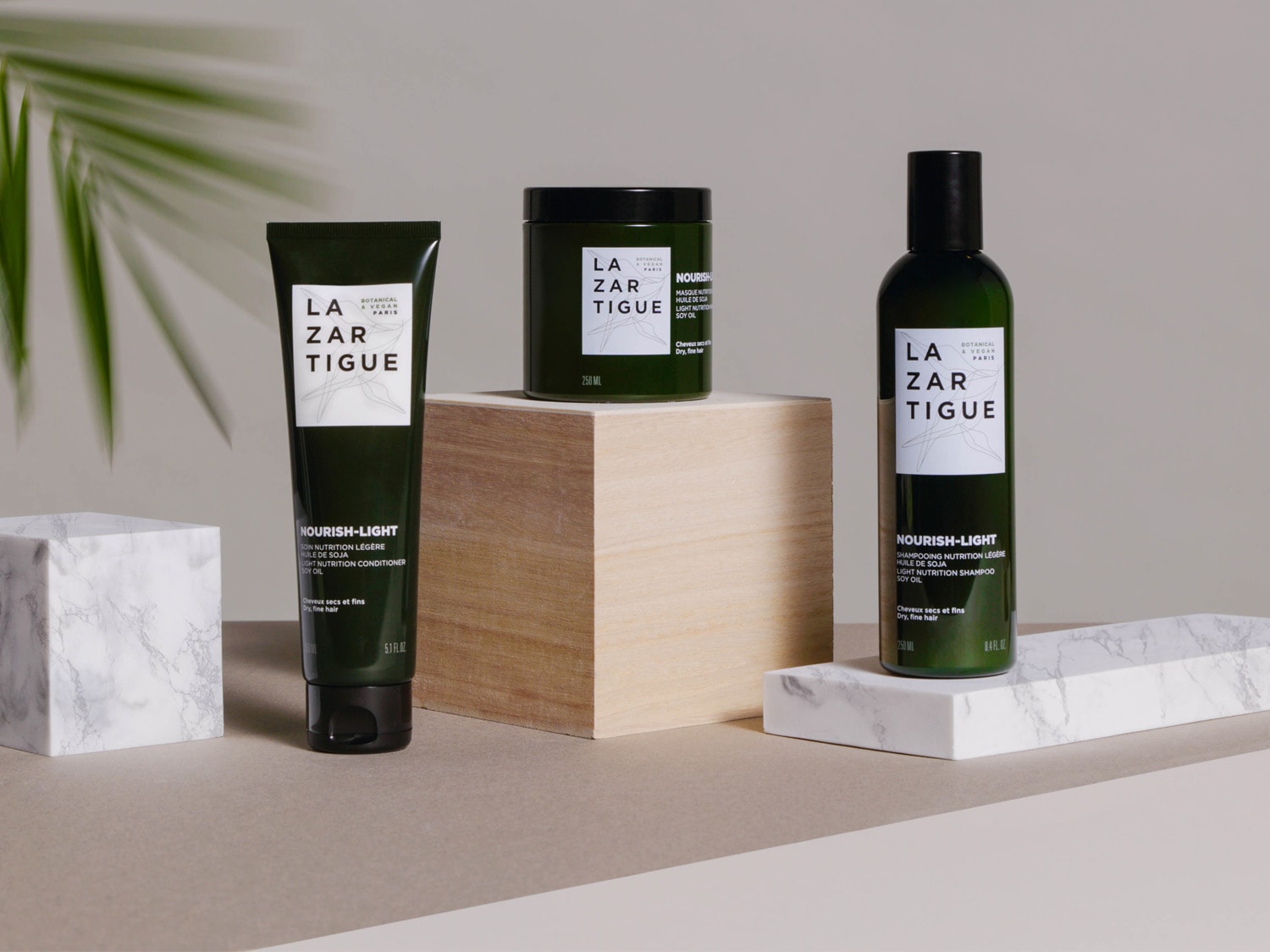 Na zdjęciu znajdują się trzy produkty do pielęgnacji włosów marki Lazartigue: odżywka, maska i szampon z linii Nourish-Light, wszystkie w ciemnozielonych opakowaniach z białymi etykietami.