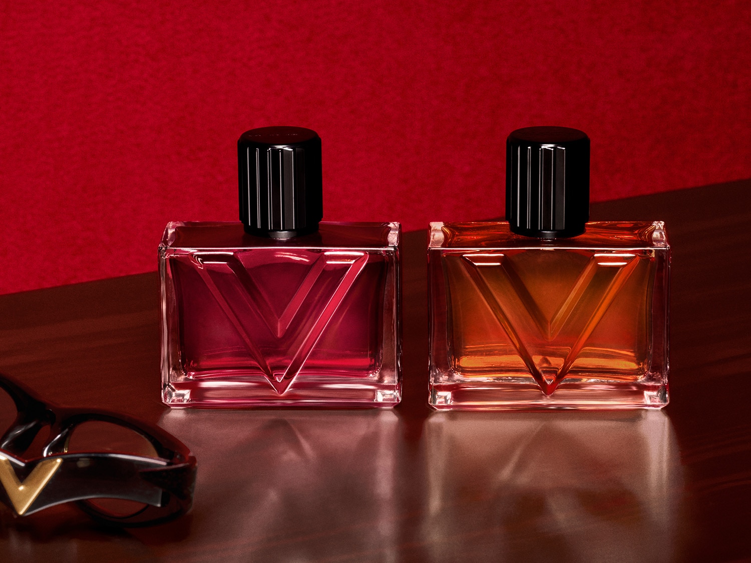Dwa prostokątne flakony perfum z czarnymi zakrętkami, jeden z czerwonawym płynem, drugi z bursztynowym, stoją na błyszczącej, brązowej powierzchni, a w tle widnieje czerwona ściana.