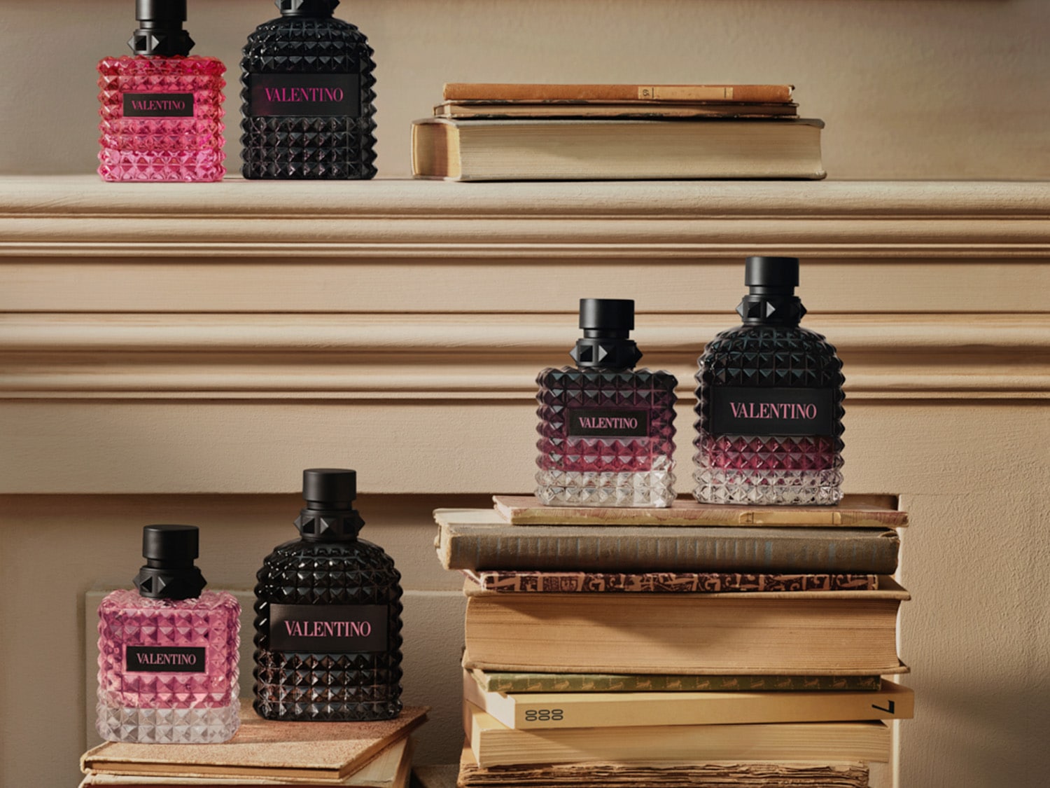 Trzy zestawy perfum Valentino, każdy składający się z dwóch flakonów o różnej wielkości, w kolorach różowym i czarnym, z ćwiekowanym wzorem, stoją na stosach książek, tworząc elegancką kompozycję.