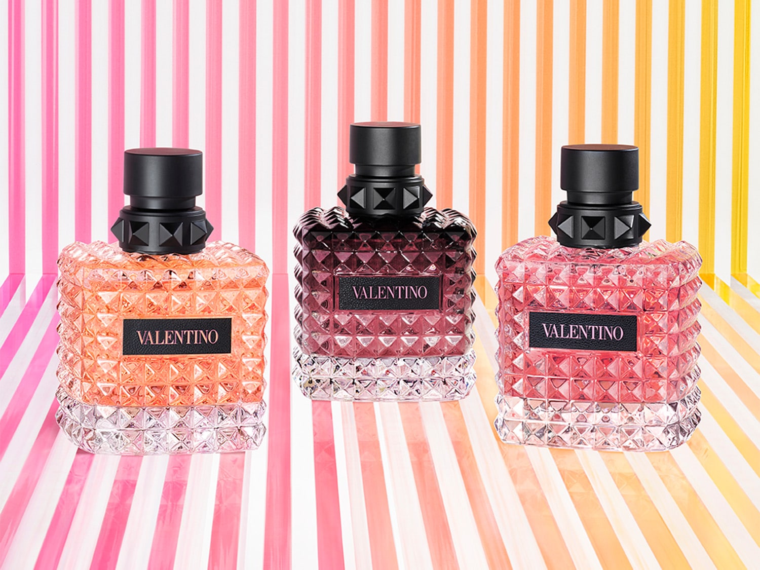 Trzy flakony perfum Valentino w odcieniach różu i pomarańczy, każdy z czarnym korkiem i nazwą marki na etykiecie, na tle kolorowych pasków.