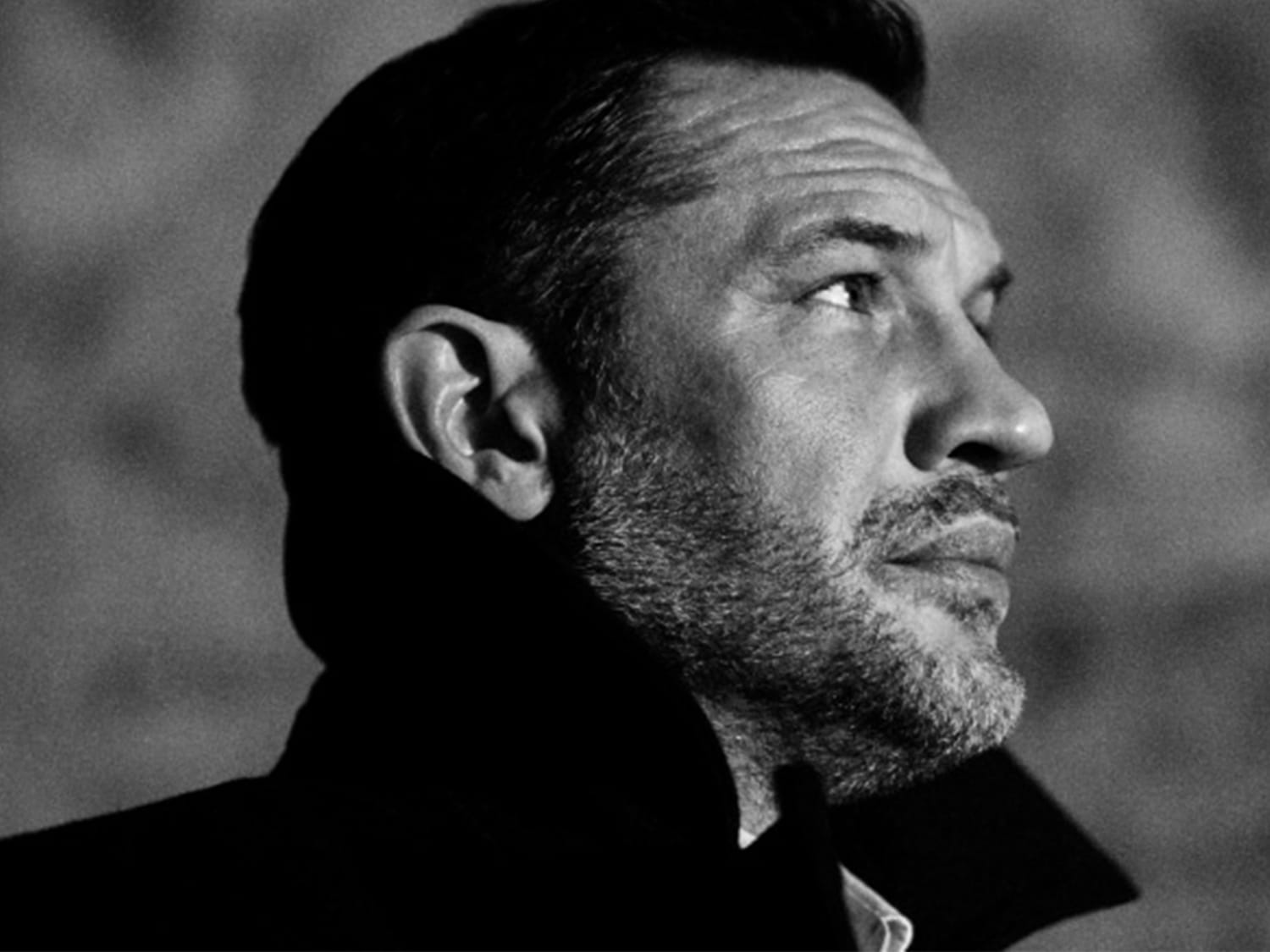 Een man met een donkere huidskleur staat voor een zandkleurige muur, gehuld in een donkerblauw gewaad, met bladeren op de voorgrond.